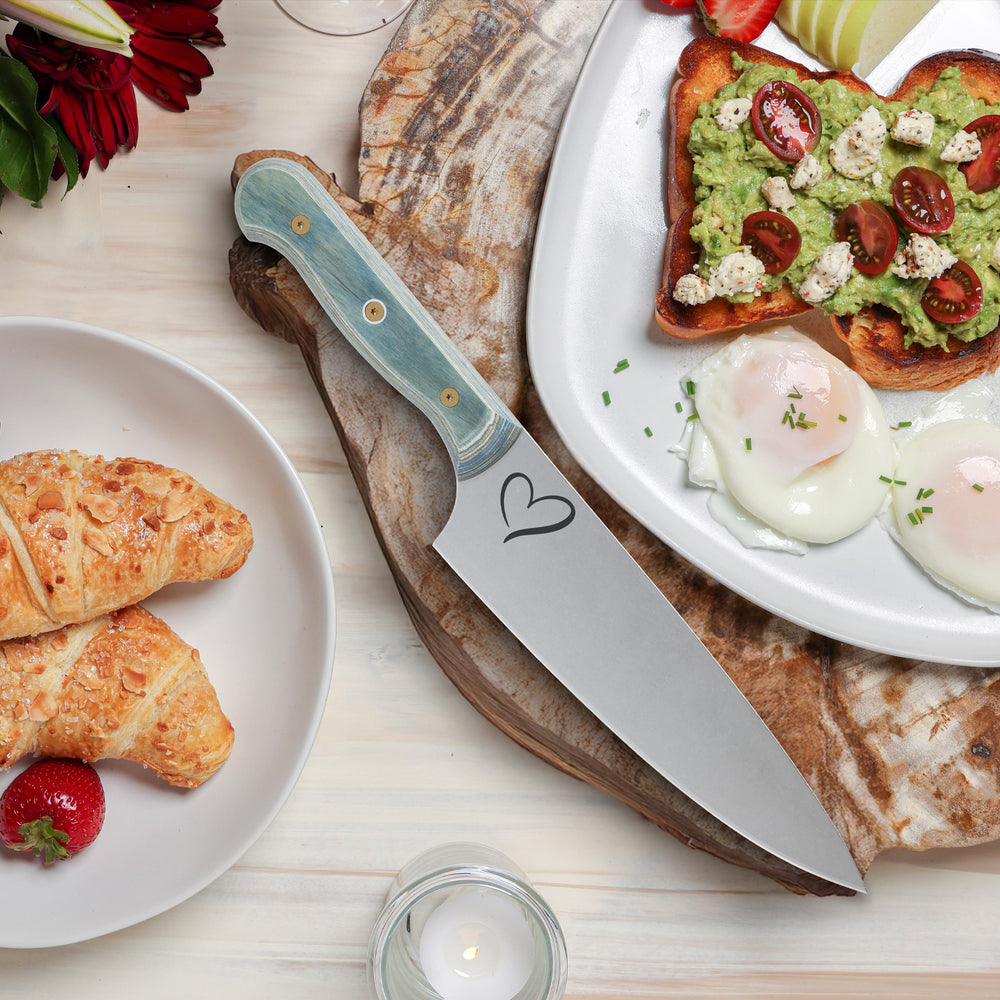 A Custom Blue Chefs Knife with a heart logo laying on wood board with eggs, avocado toast and croissants.