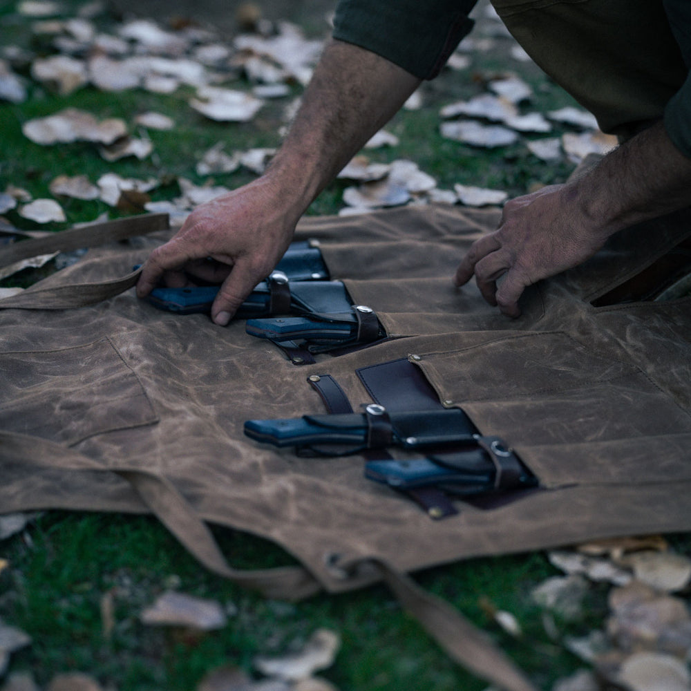 Person arranging knives in leather sheaths in a brown leather knife roll outdoors.