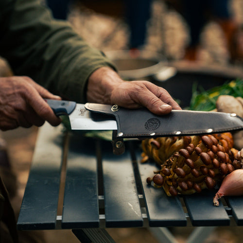 Chef Adam Glick holding a knife that has a leather sheath on it over mushroom on a table