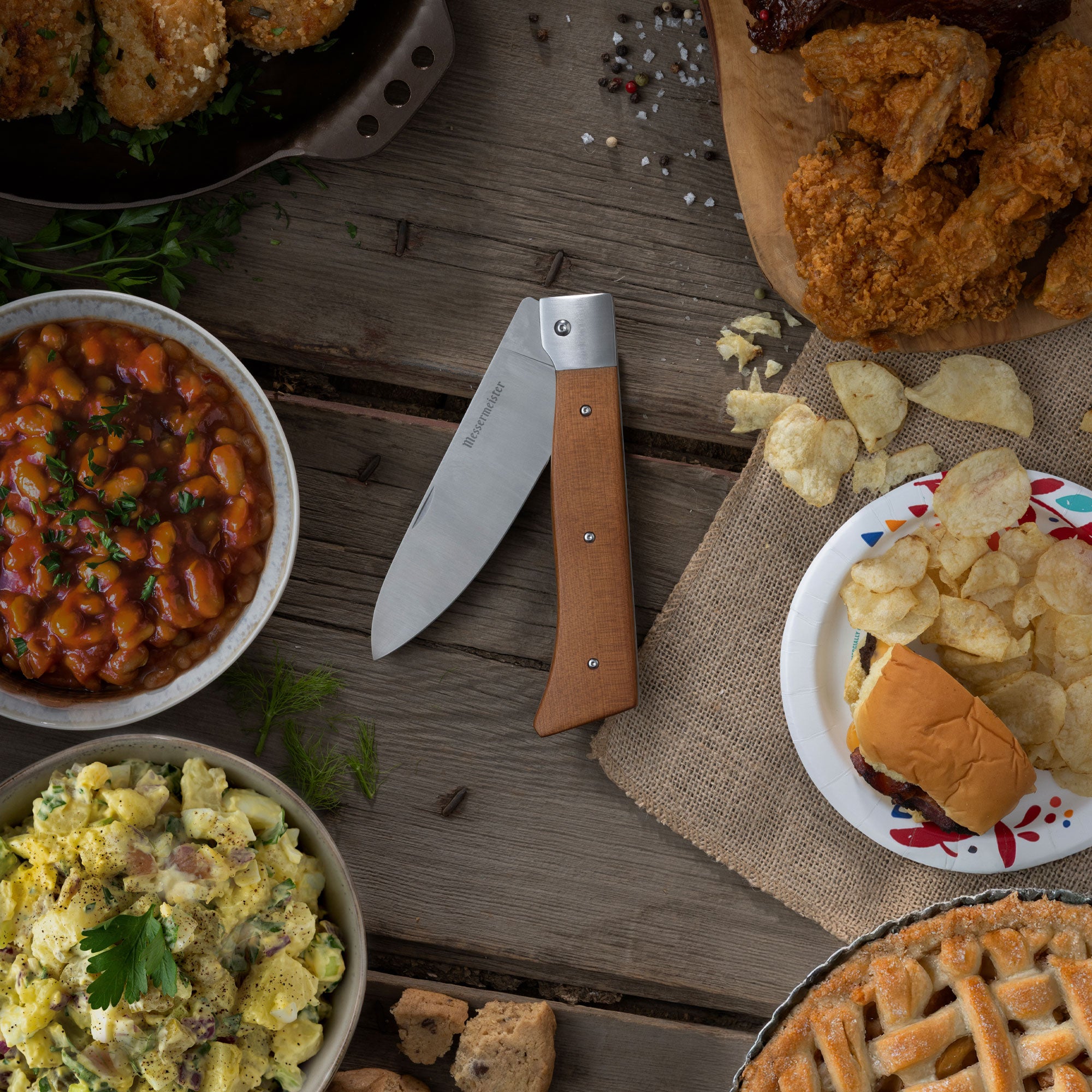 Messermeister Adventure Chef Folding Chef Knife pictured at a picnic table with Beans, Potato Salad, Chips, Apple Pie, Crab Cakes and Fried Chicken