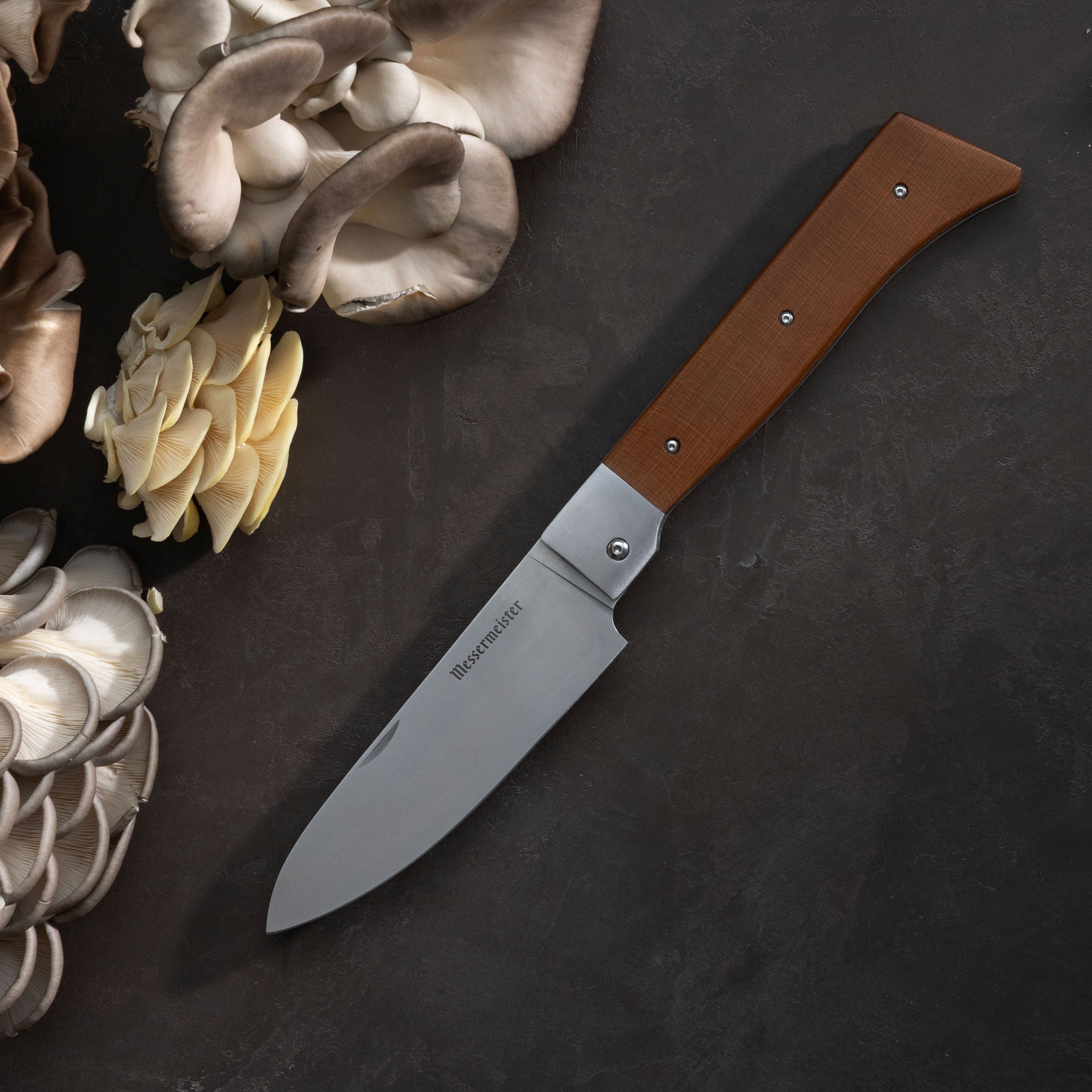 Messermeister Adventure Chef Folding Chef's Knife with a wooden handle on a dark surface with mushrooms