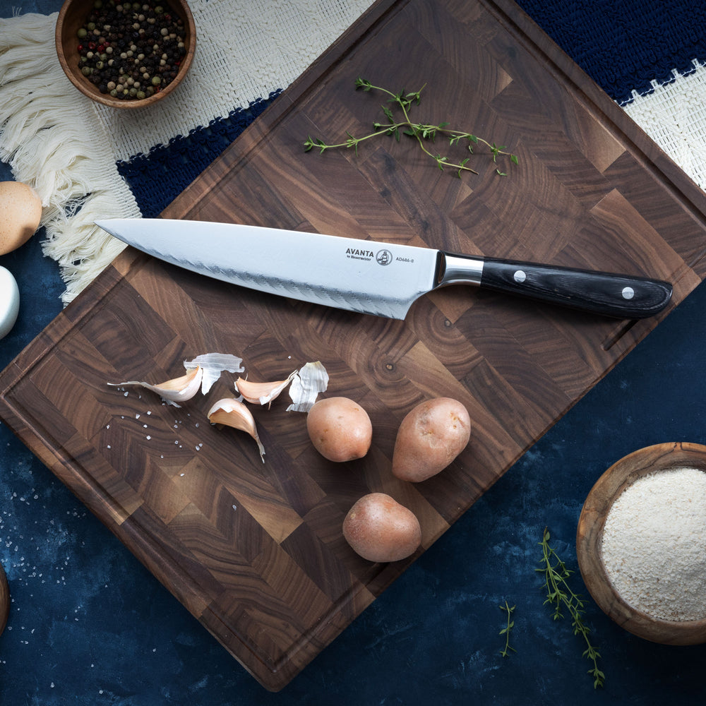 Wooden cutting board with a Messermeister Avanta Damascus Chef's Knife, garlic, and potatoes on a dark blue surface