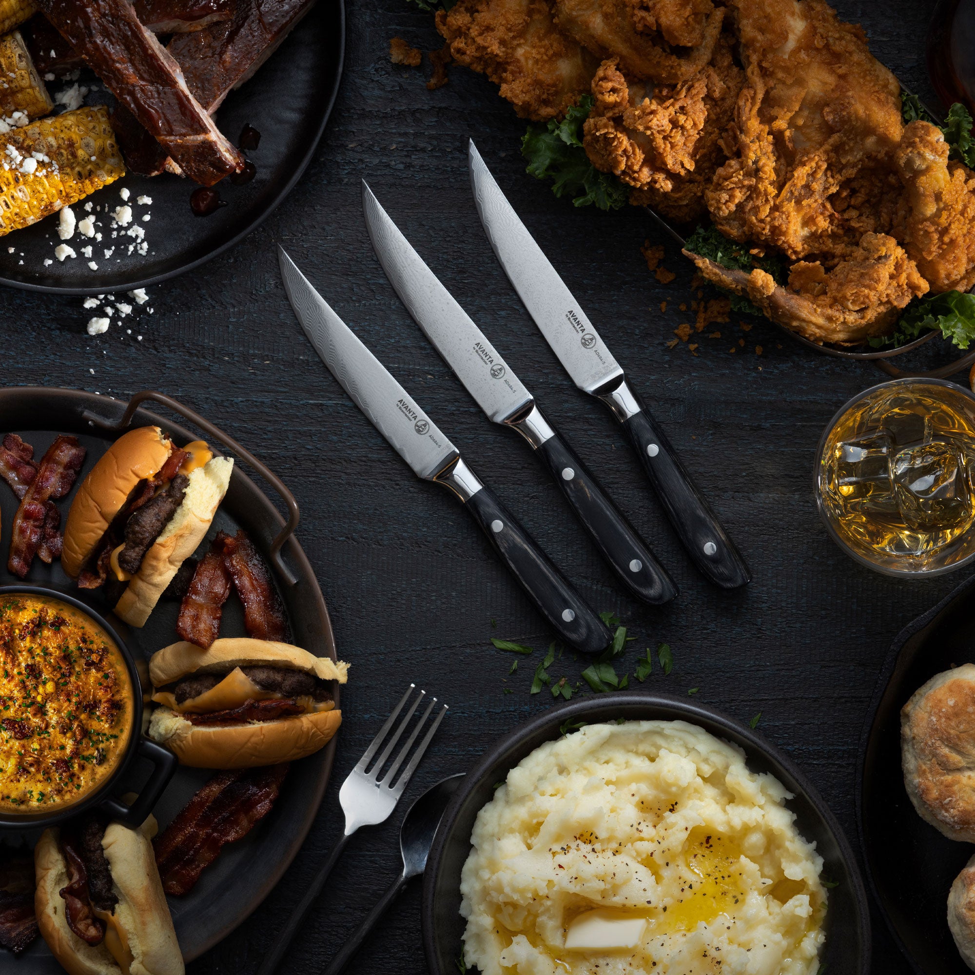 Assorted food items including burgers, fries, and mashed potatoes with Messermeister Avanta Damascus Steak Knives on a dark surface.