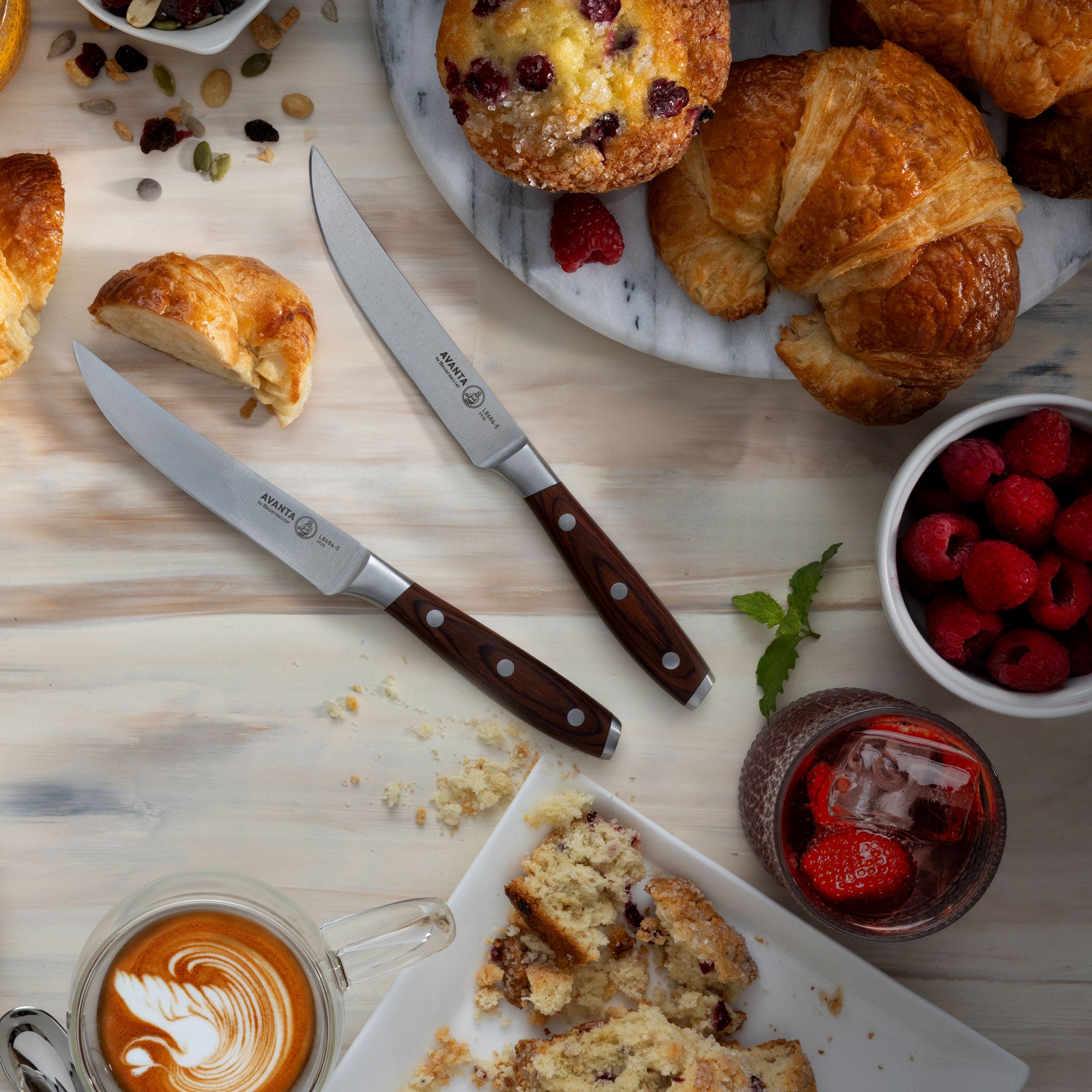 Assorted pastries on a wooden table with 2 Messermeister Avanta Pakkawood Steak Knives and drinks.