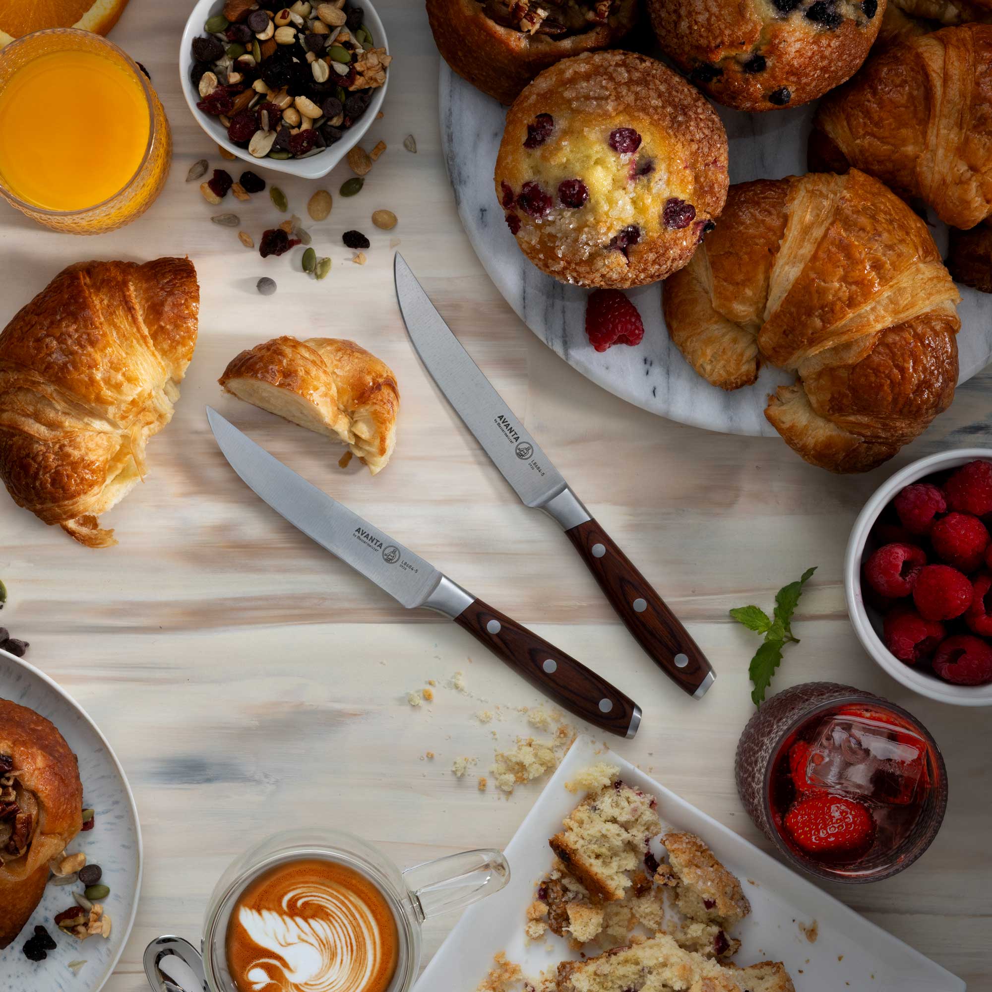 Breakfast scene with pastries, fruit, and coffee on a white wash wooden table with 2 Messermeister Avanta Pakkawood Steak Knives.