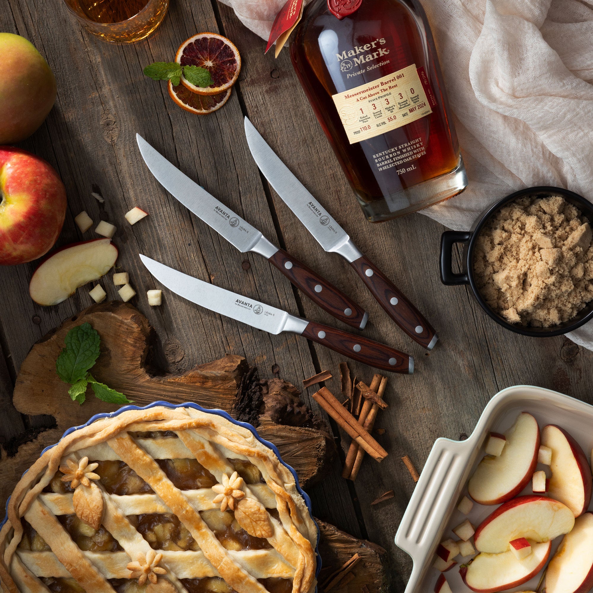 Wooden table with an apple pie, 3 Messermeister Avanta Pakkawood Steak Knives, apples, and a bottle of Maker's Mark bourbon.