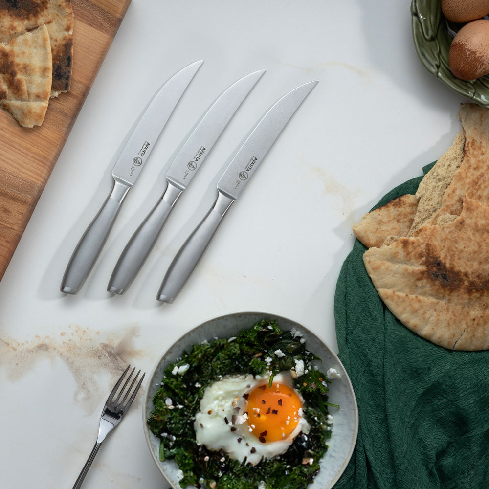 Three Messermeister Avanta Stainless Steel Steak Knives on a marble surface with a bowl with a cooked egg and greens with pita bread.