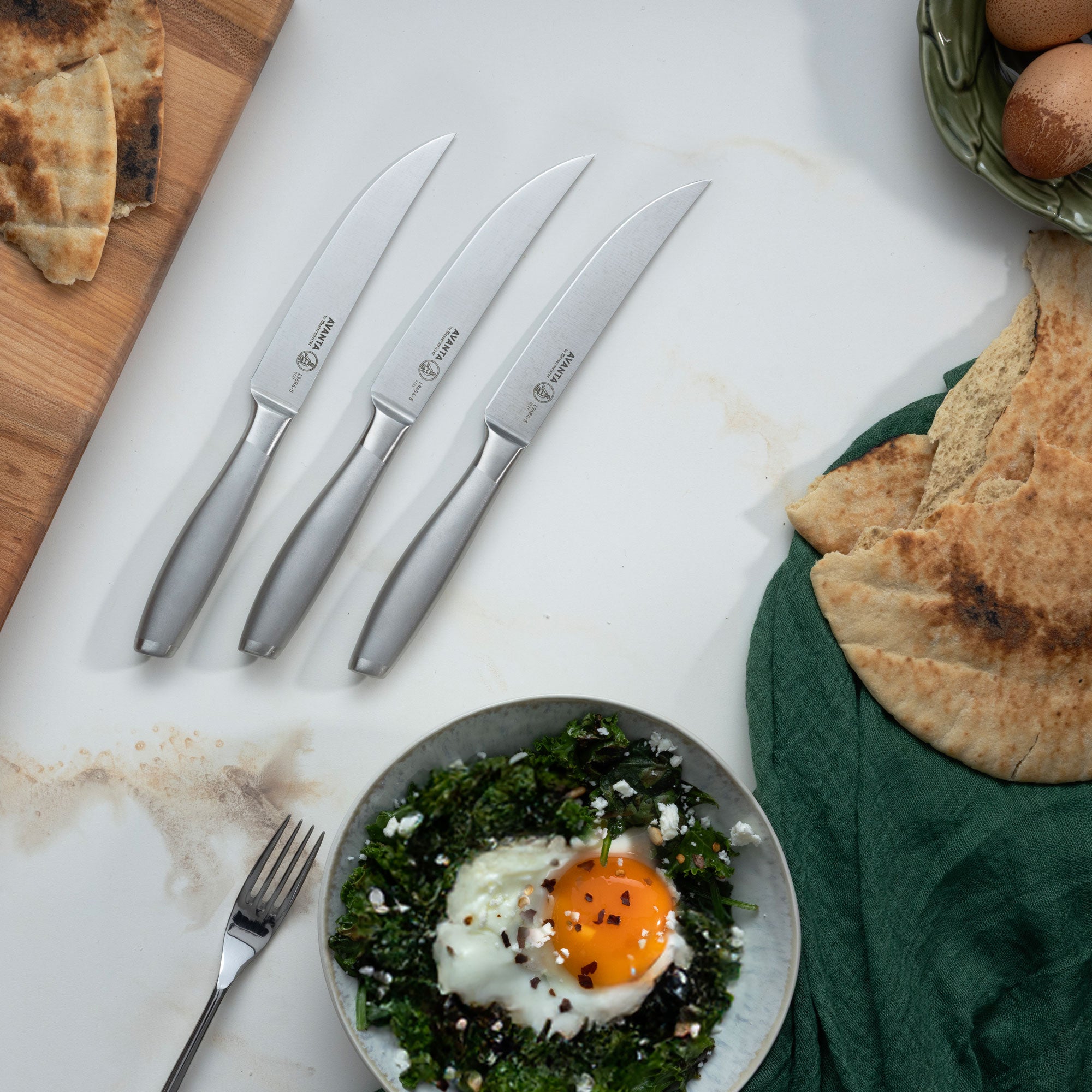 Three Messermeister Avanta Stainless Steel Steak Knives on a marble surface with a bowl with a cooked egg and greens with pita bread.