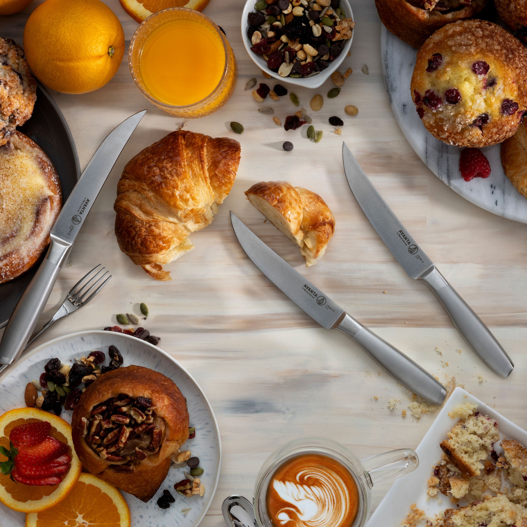 Breakfast scene with pastries, coffee, and 3 Messermeister Avanta Stainless Steel Steak Knives on a wooden surface