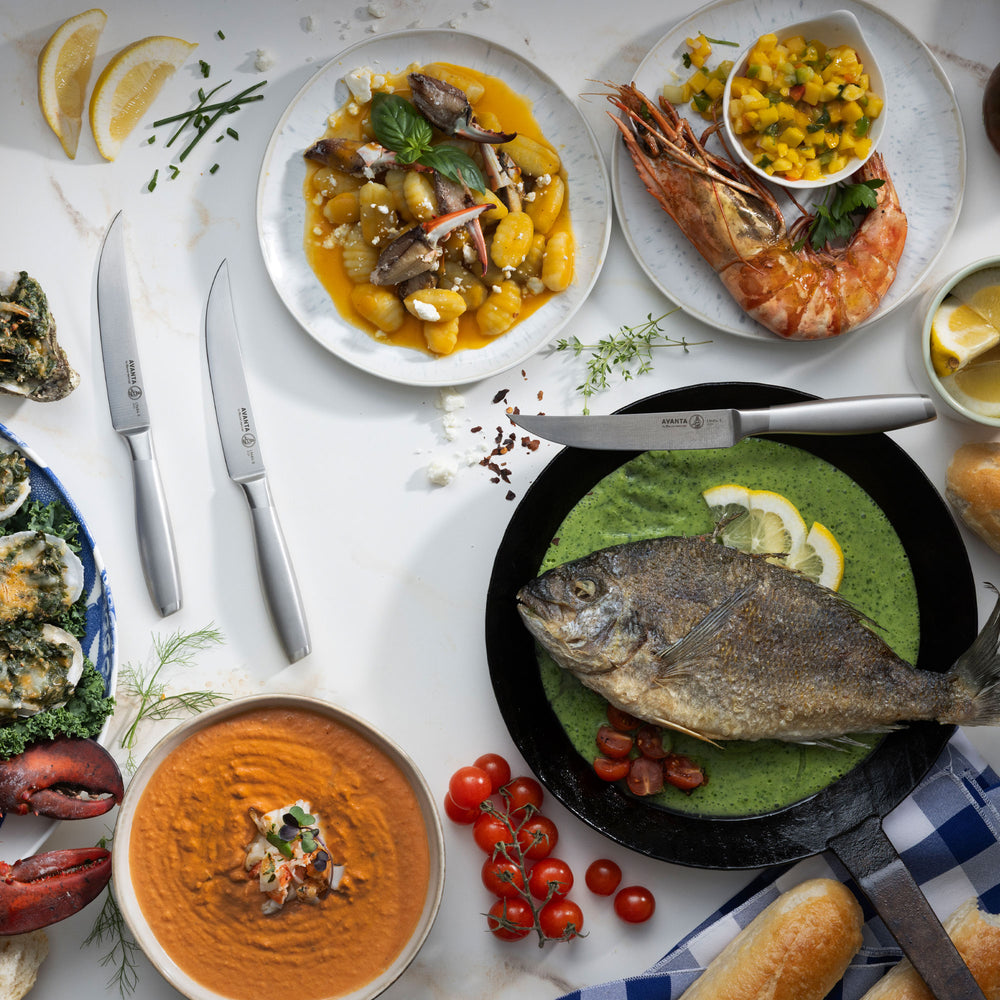 Assorted dishes including a fish dish, prawns, Messermeister Avanta Stainless Steel Steak Knives, and hummus on a white table.