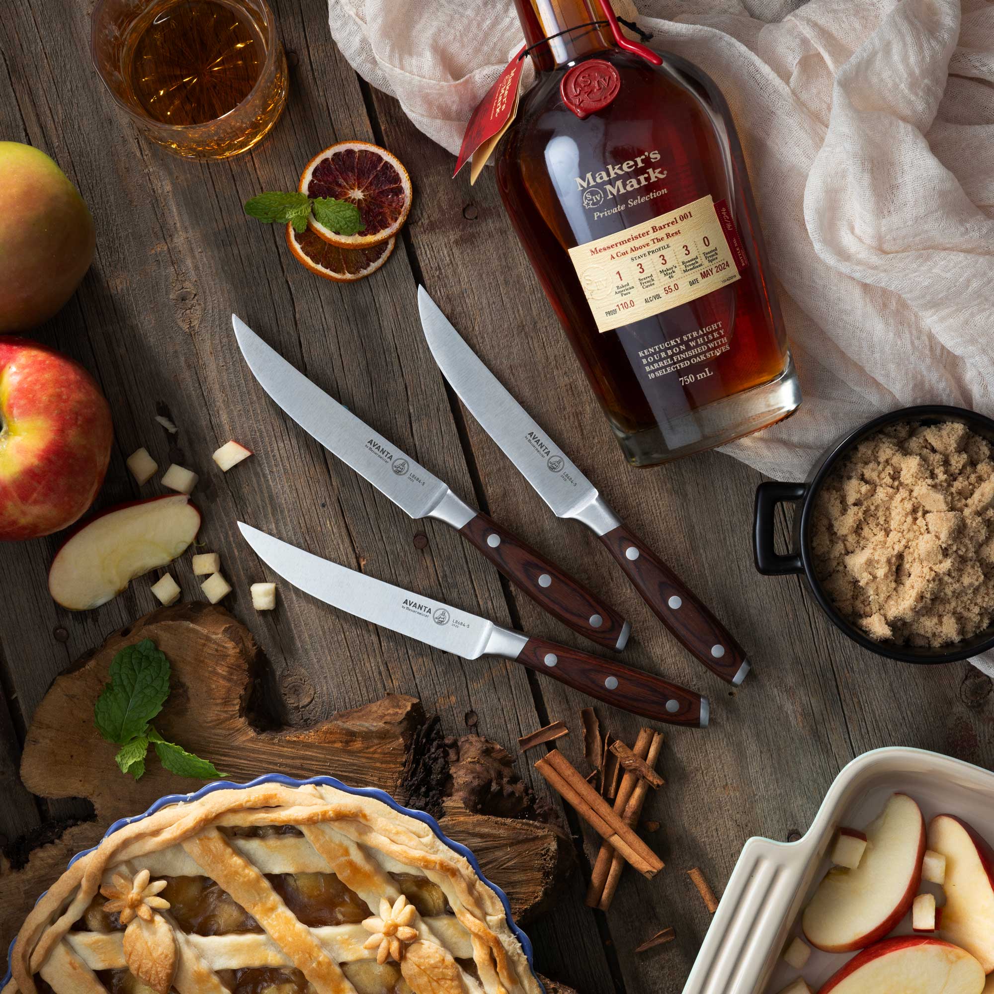 Bottle of Maker's Mark bourbon with 3 Messermeister's Avanta Steak Knives, apples, and a pie on a wooden surface.