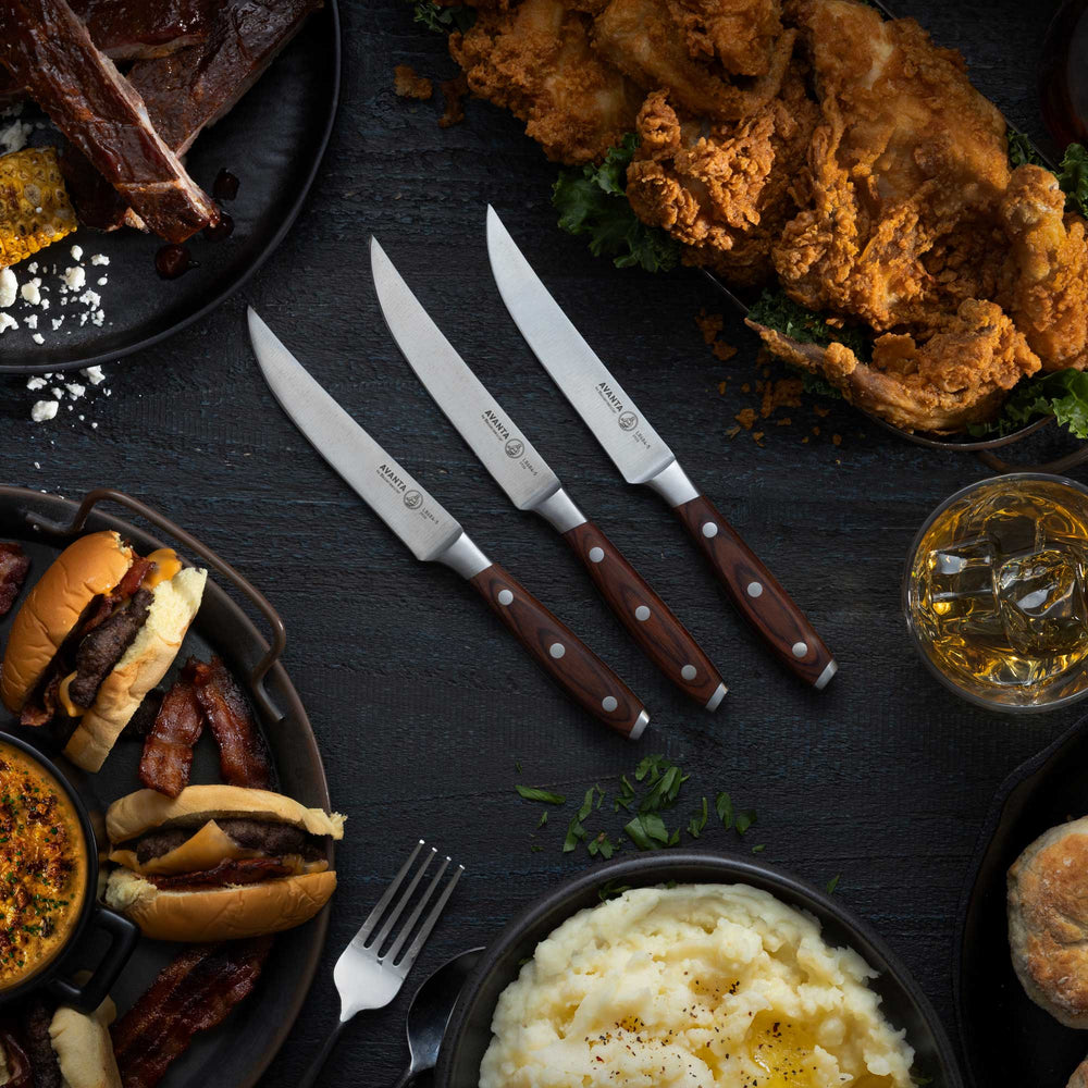 Assorted food items including burgers, fried chicken, mashed potatoes, ribs, and a glass of beer on a dark surface with 3 Messermeister Avanta Pakkawood Steak Knives.