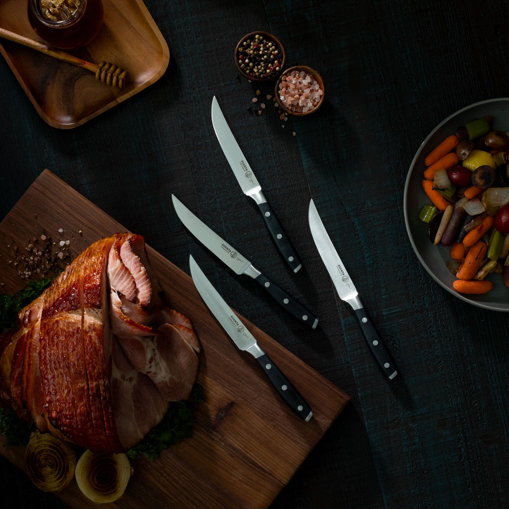 Messermeister Avanta Steak Knives with Black Handles pictured on dark suraface with Spiral Sliced Honey Glazed Ham Dinner and Roasted Vegetable