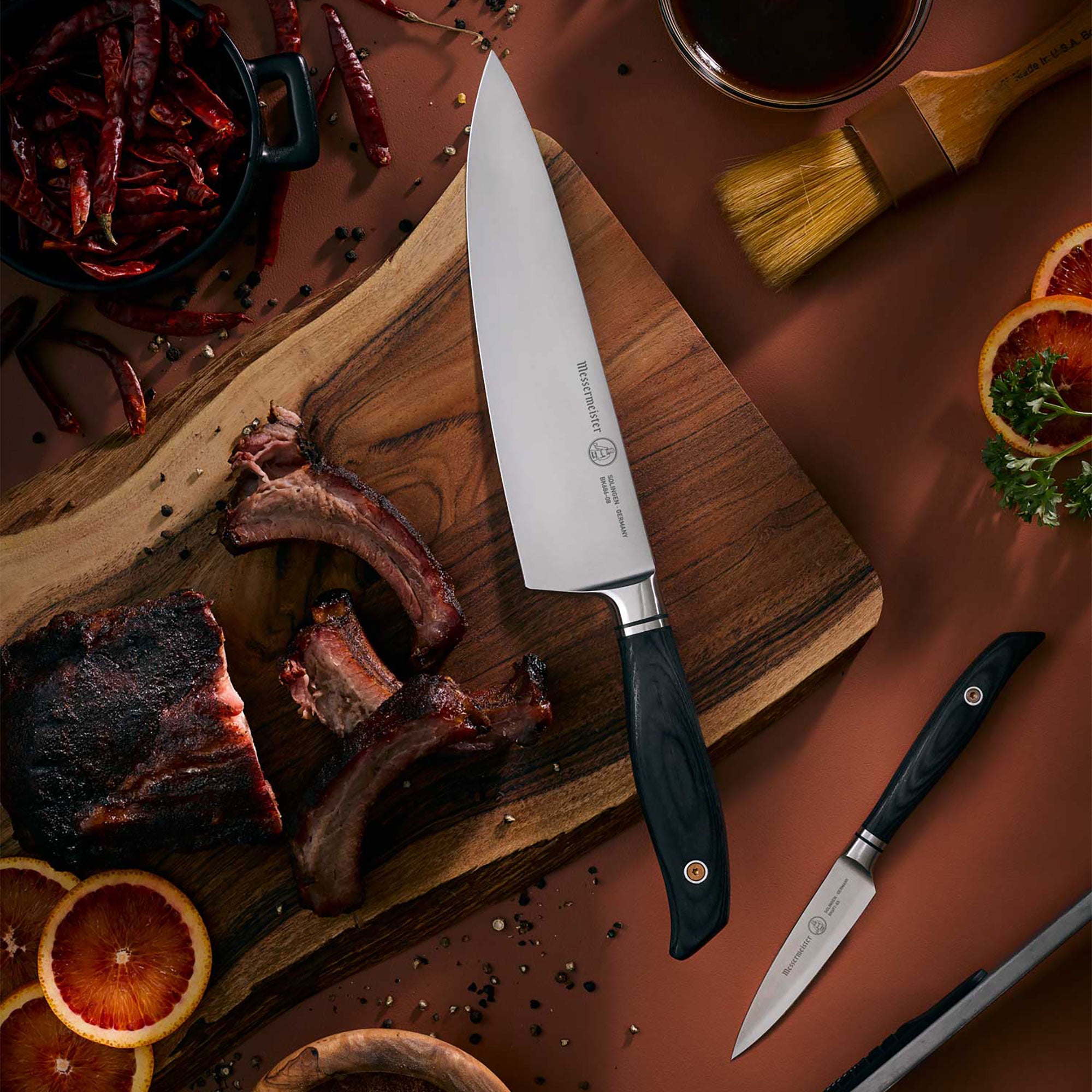 Messermeister Blacksmith Chef Knife and Paring Knife on Cutting Board with Ribs and spices on dark surface and sliced Oranges