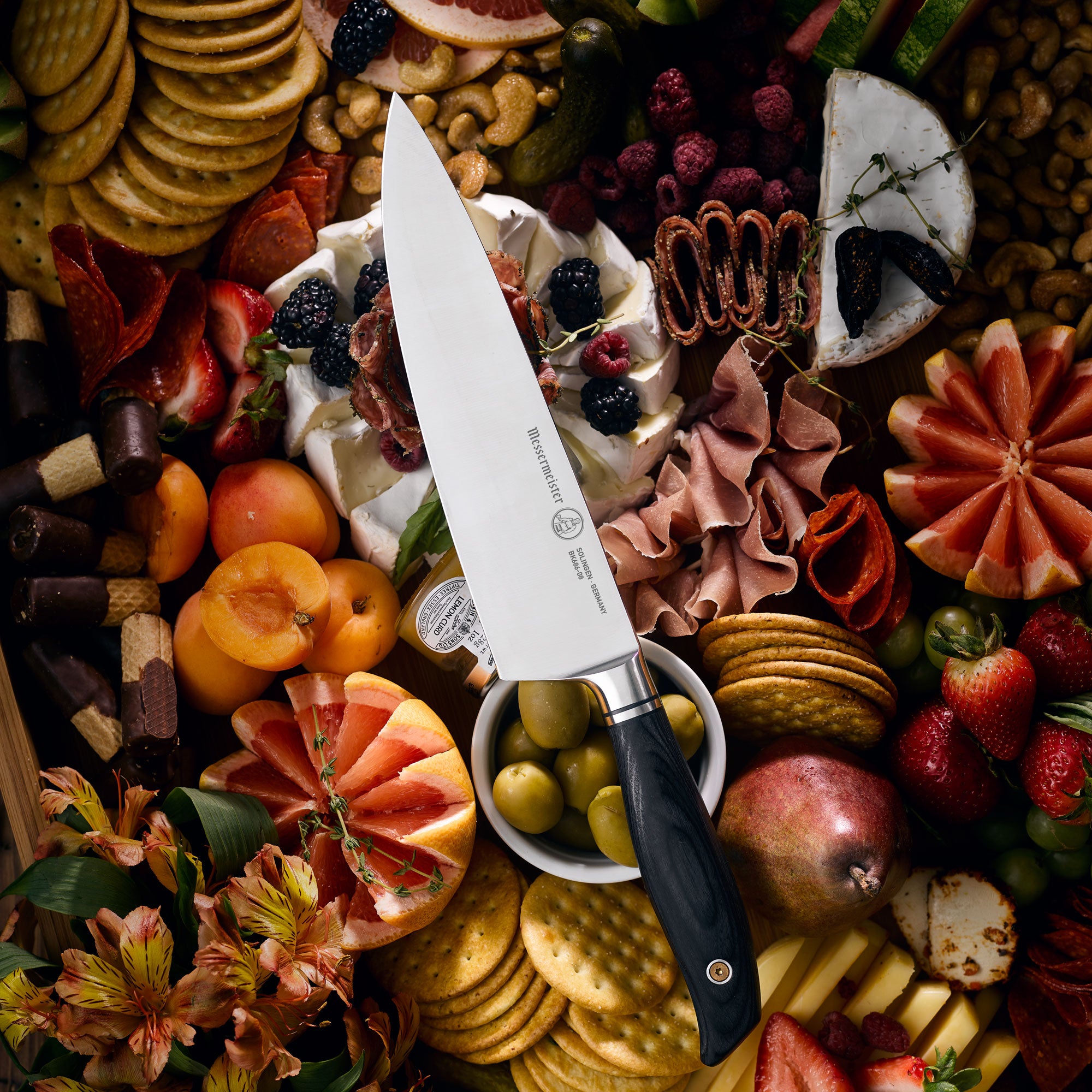 Messermeister Blacksmith Chef Knife resting on a charcuterie board surrounded by sliced cured meats, artisan cheeses,  crackers, fresh herbs, and seasonal fruits, highlighting the knife’s satin-finish blade and black handle