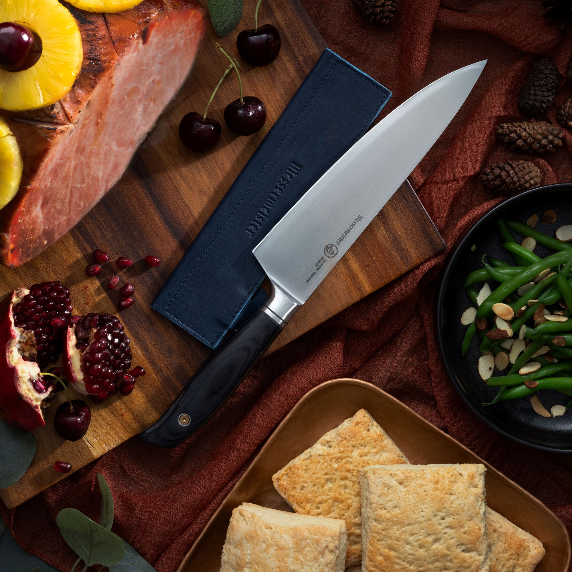 Messermeister Blacksmith Chef's Knife and a Leather Magnetic Edge-Guard on a wooden cutting board with food items like ham, pomegranate, and green beans.