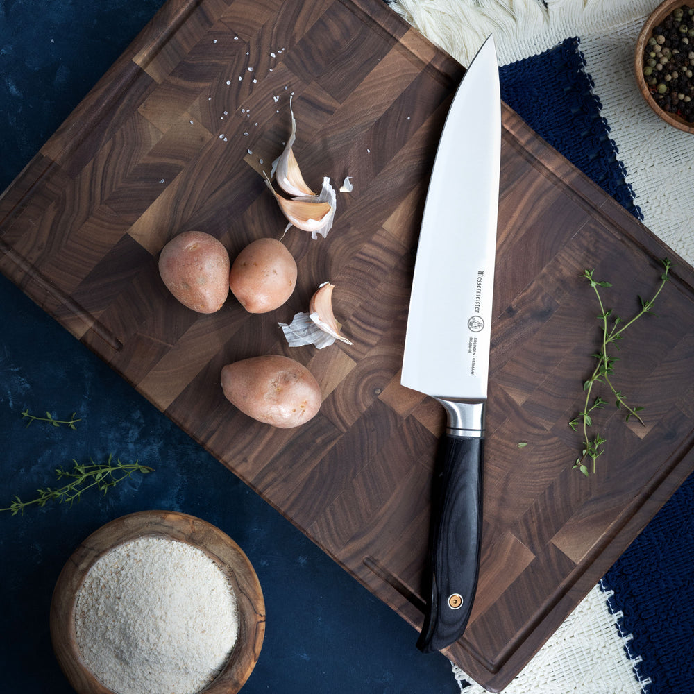 Wooden cutting board with a Messermeister Blacksmith Chef Knife, potatoes, garlic, and herbs on a dark blue surface.