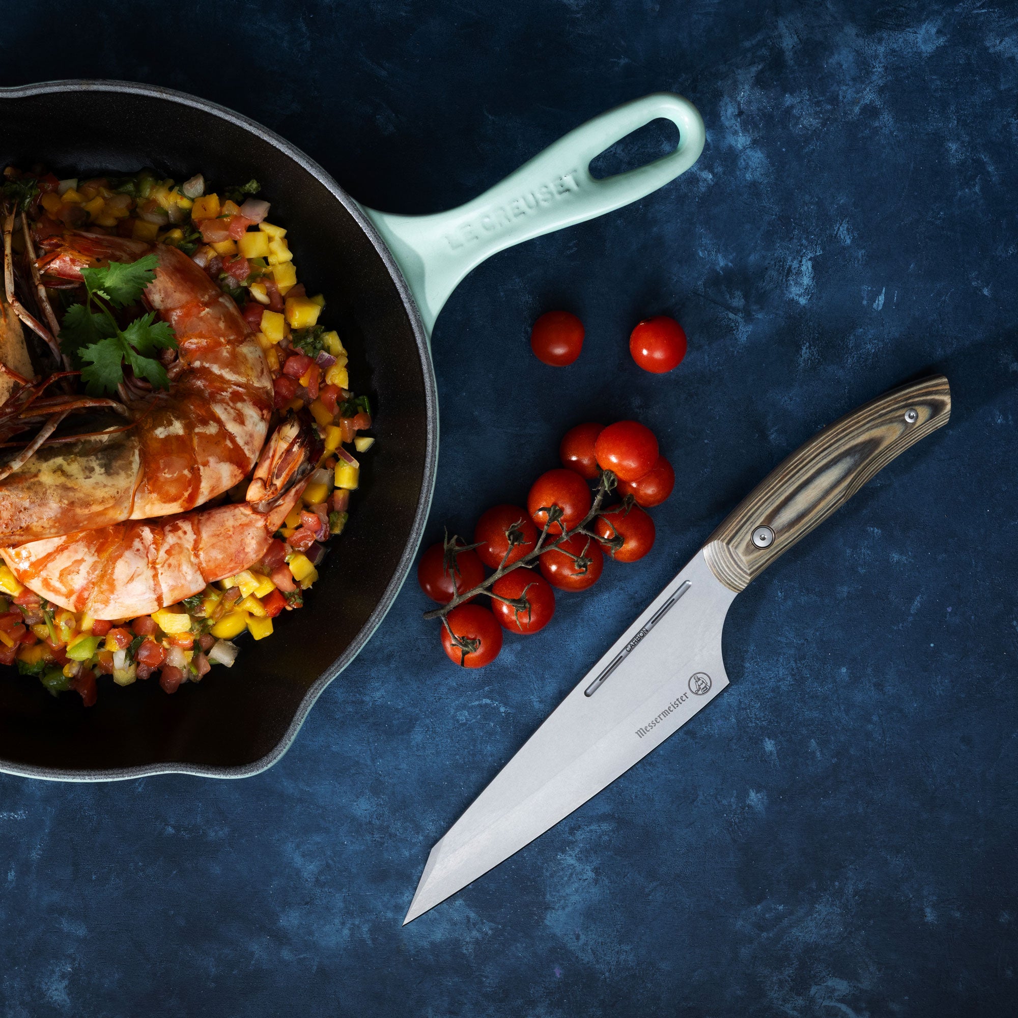 Skillet with shrimp and vegetables on a dark surface with a Messermeister Carbon April Bloomfield Chef Knife and cherry tomatoes.