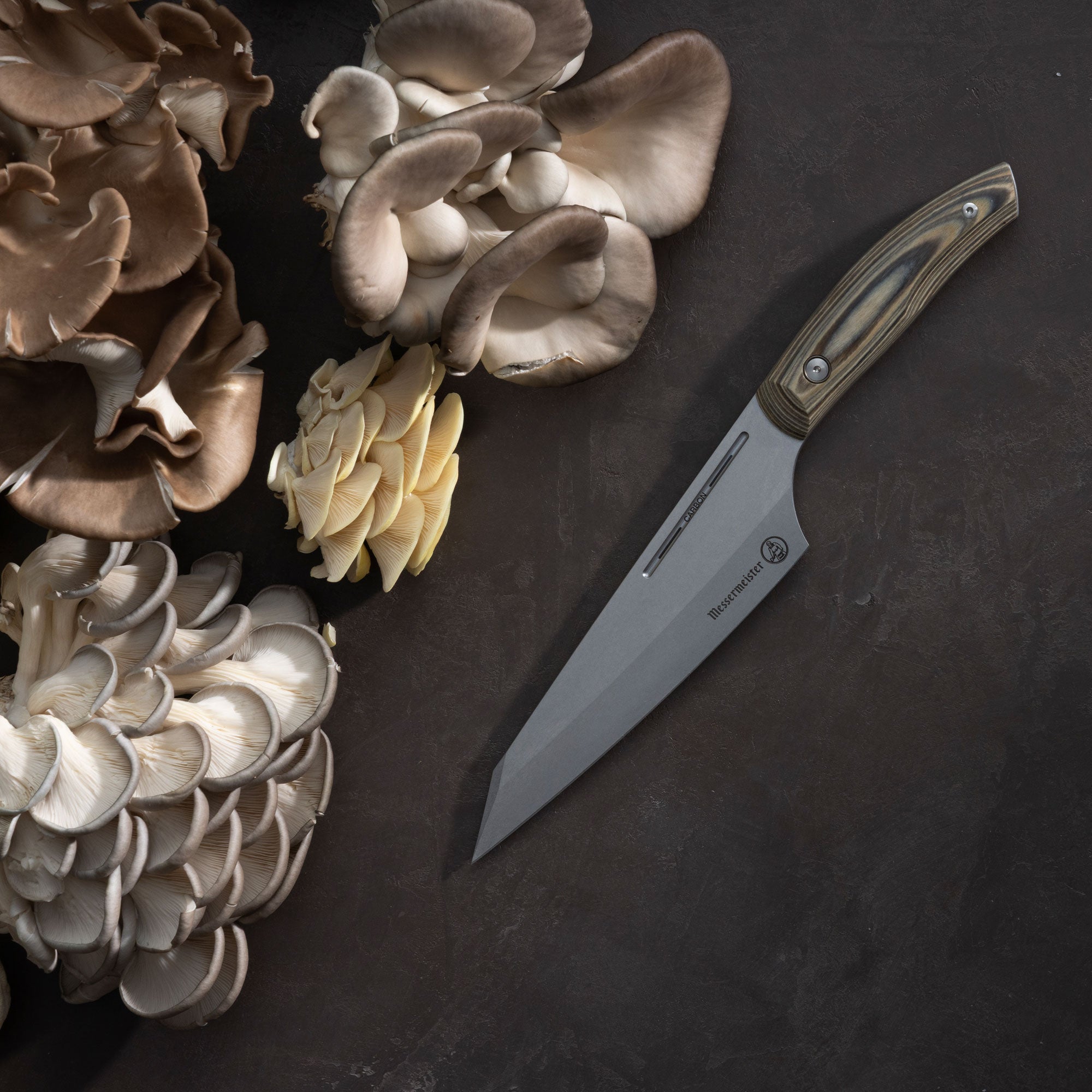 Messermeister Carbon April Bloomfield Chef Knife next to various types of mushrooms on a dark surface