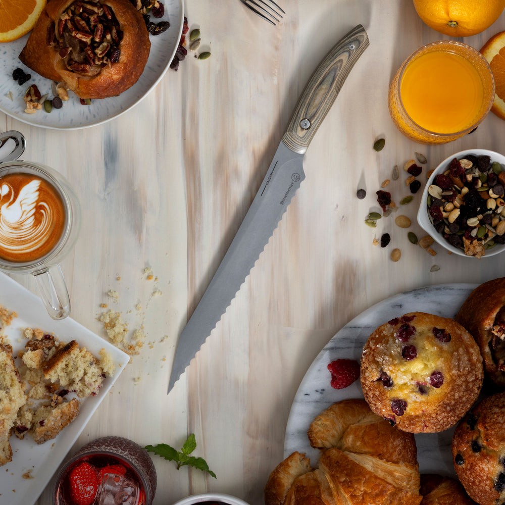 Messermeister Carbon Bread Knife with a breakfast spread with pastries, coffee, and fruit on a wooden surface.