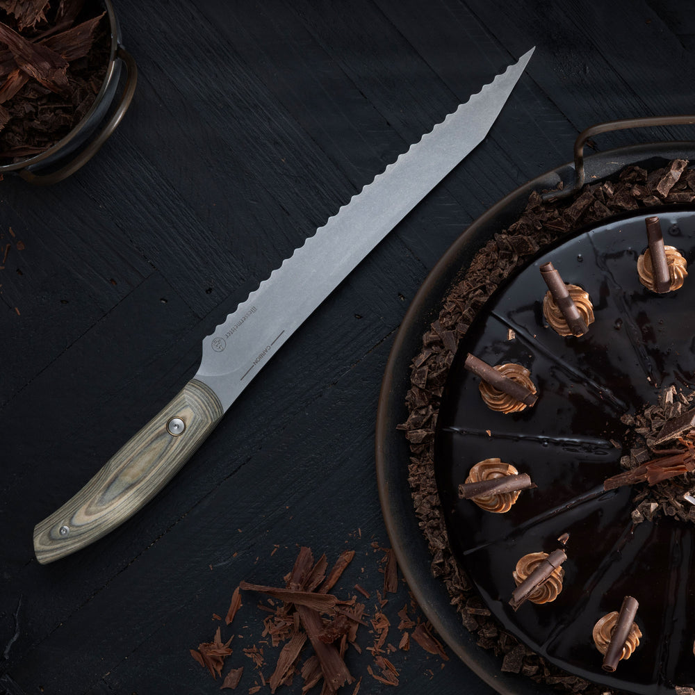 Messermeister Carbon Scalloped Slicer laying besides a dark chocolate cake on a black wood surface.