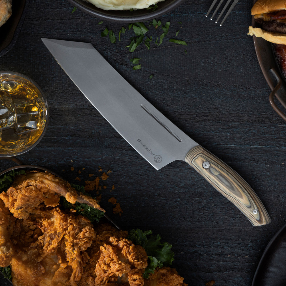 Messermeister Carbon Bunka Knife on a dark surface with fried chicken, a glass of bourbon, and a burger.