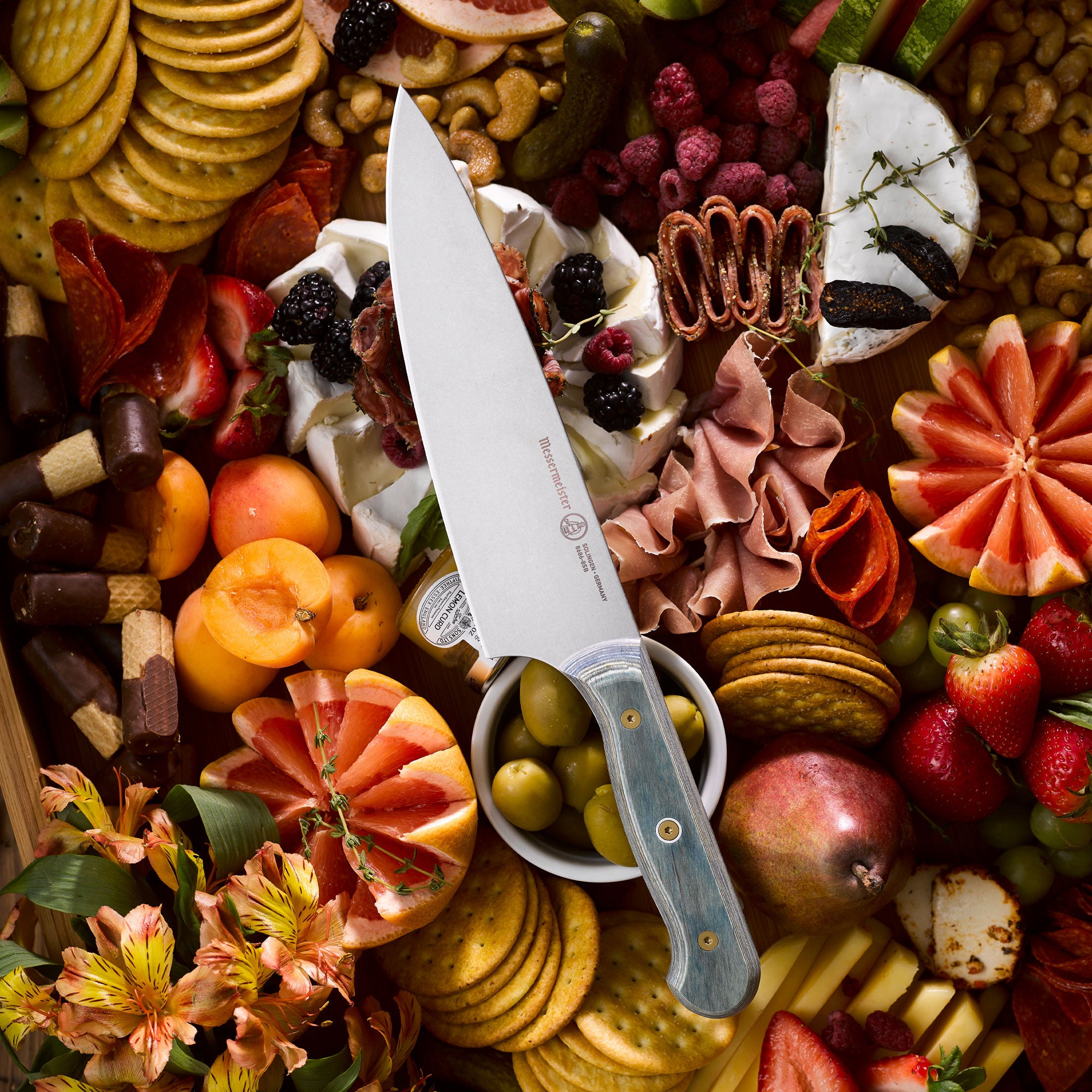 Messermeister Custom Blue Chef Knife resting on a charcuterie board surrounded by sliced cured meats, artisan cheeses,  crackers, fresh herbs, and seasonal fruits, highlighting the knife’s satin-finish blade and blue handle