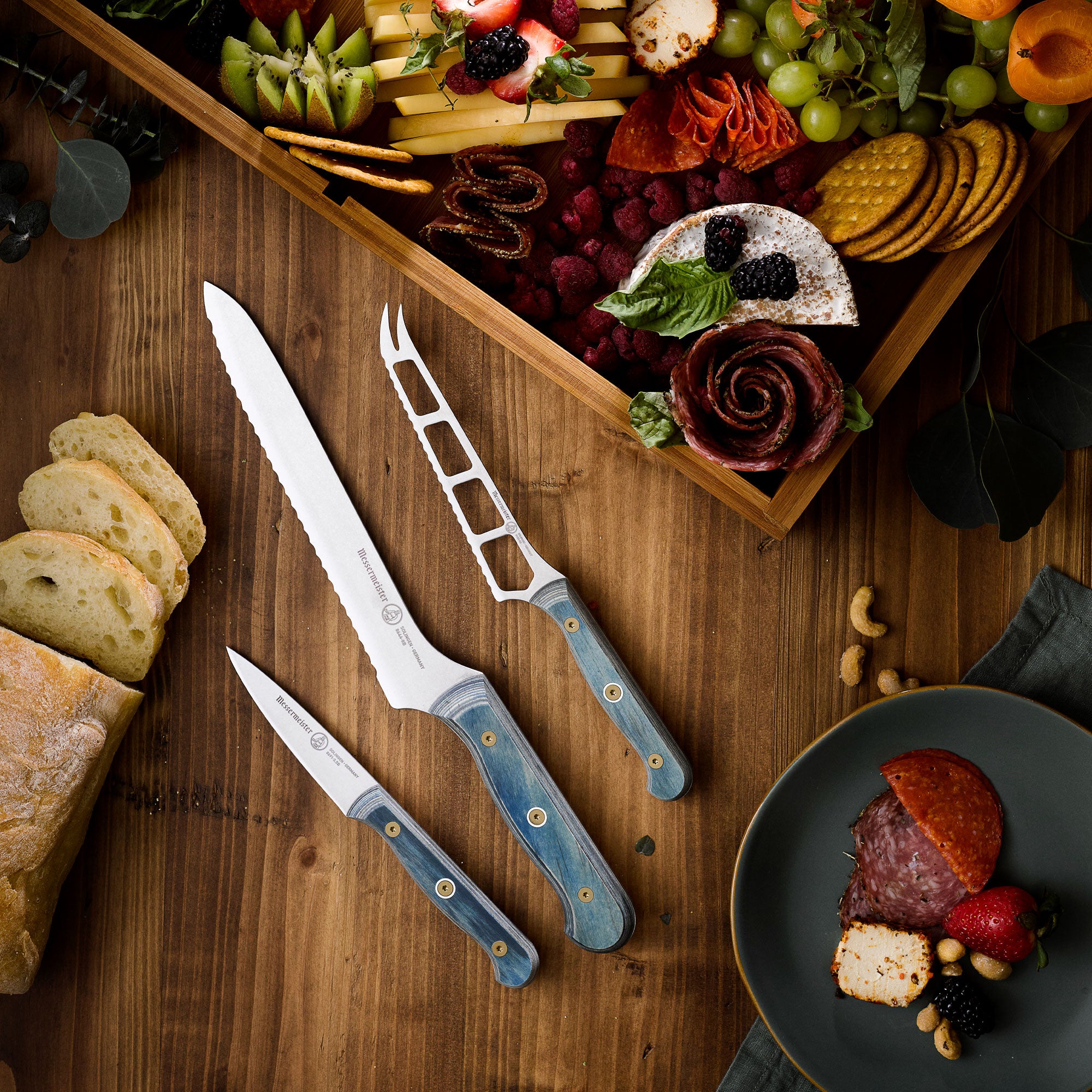 Wooden charcuterie board with assorted meats, cheeses, and bread, accompanied by a Messermeister Custom Blue Cheese & Tomato Knife, Scalloped Offset Bread Knife and Paring Knife on a wooden surface.