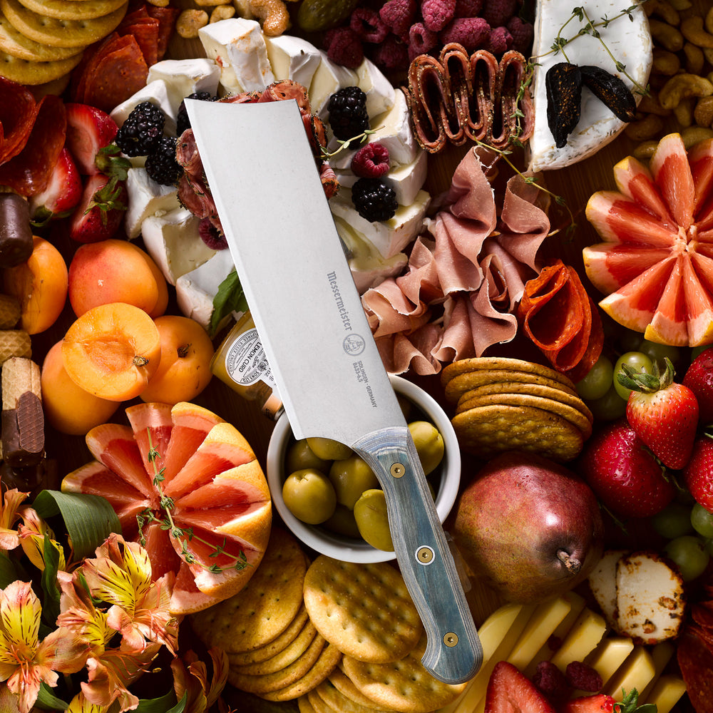 Messermeister Custom Blue Nakiri knife resting on a charcuterie board surrounded by sliced cured meats, artisan cheeses,  crackers, fresh herbs, and seasonal fruits, highlighting the knife’s satin-finish blade and blue handle