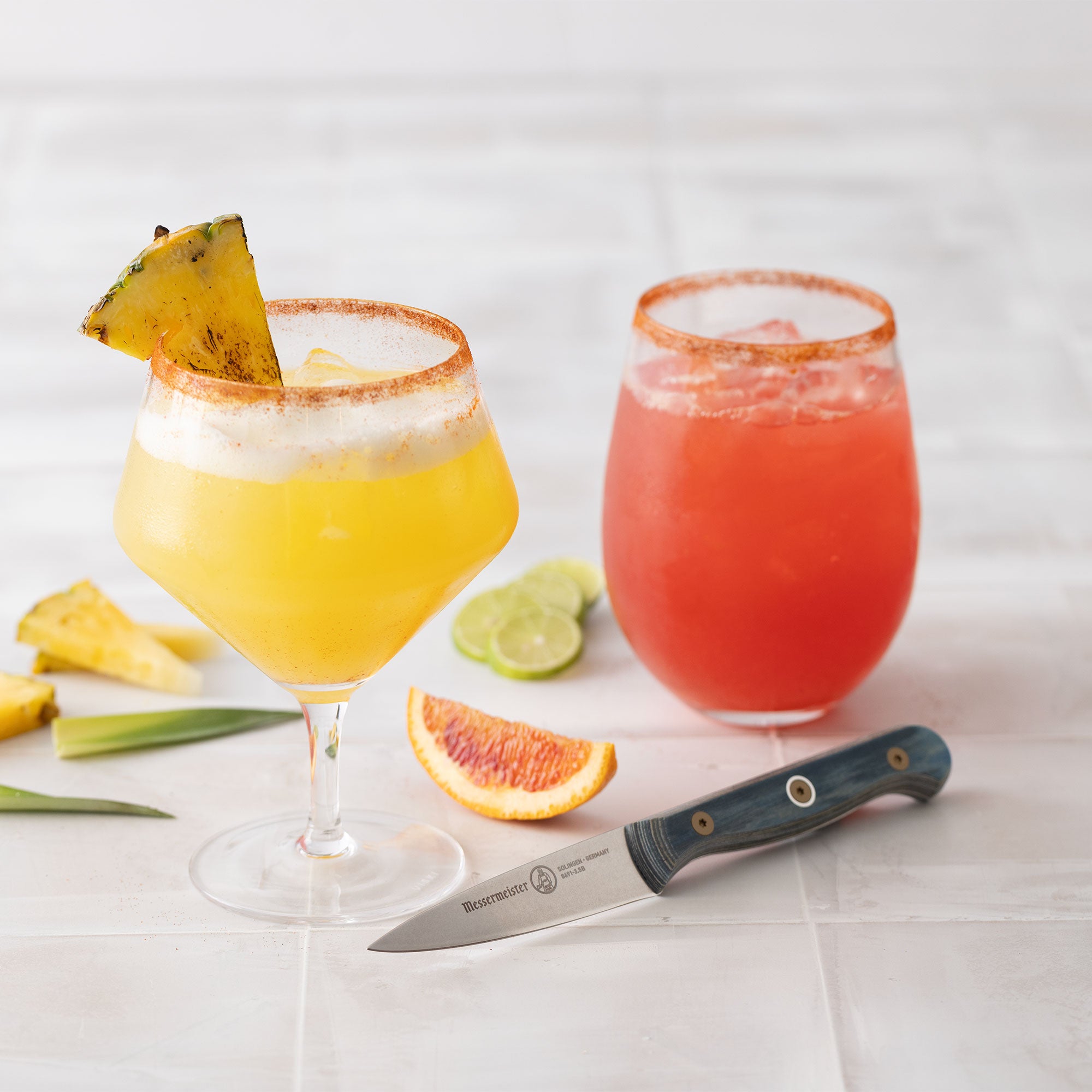 Two colorful cocktails with garnishes on a white surface, accompanied by a Messermeister Custom Blue Paring Knife