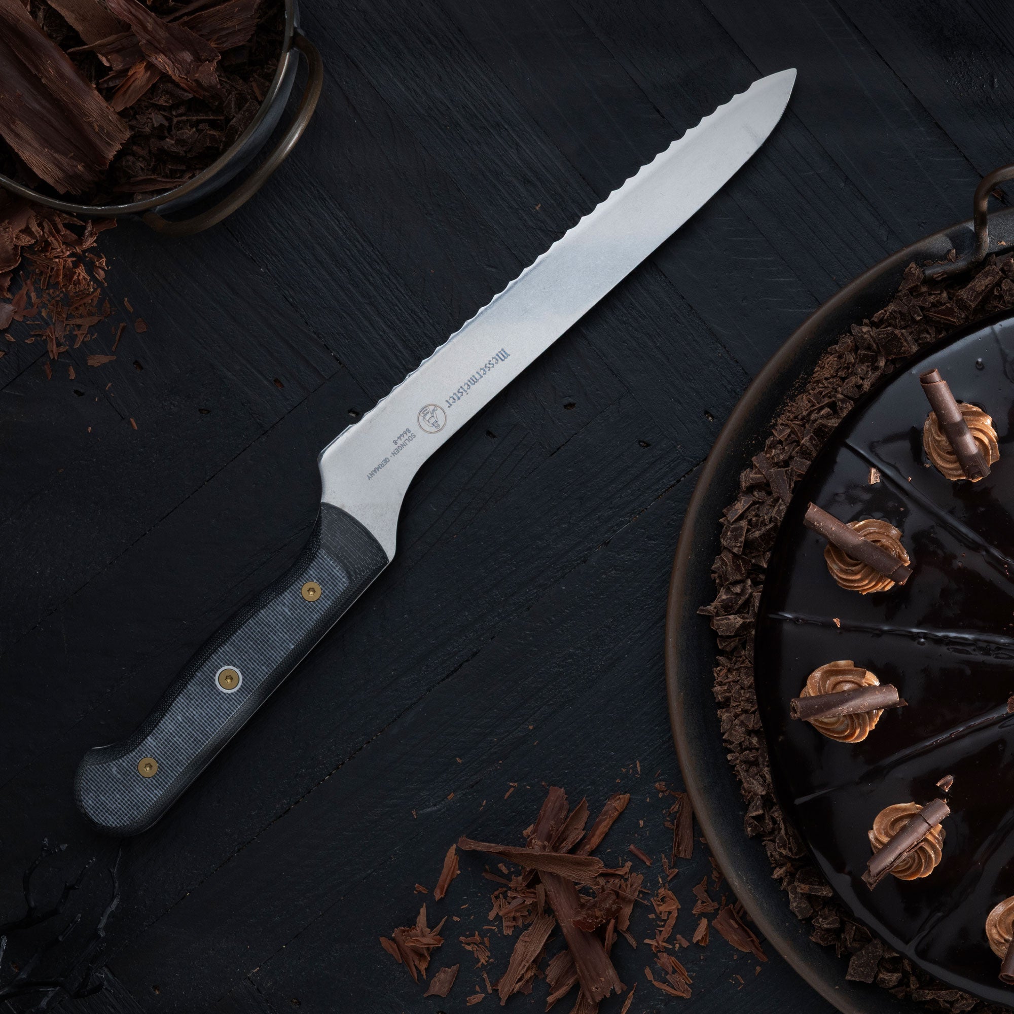 Messermeister Custom Offset Scalloped Slicer Bread Knife laying besides a dark chocolate cake on a black wood surface.