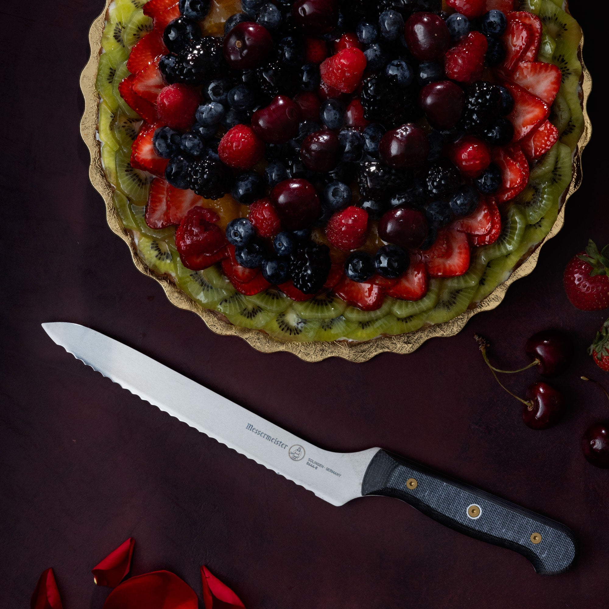 Messermeister Offset Scalloped Slicing Bread Knife with a fruit tart, cherries, strawberries and rose petals on a dark red surface