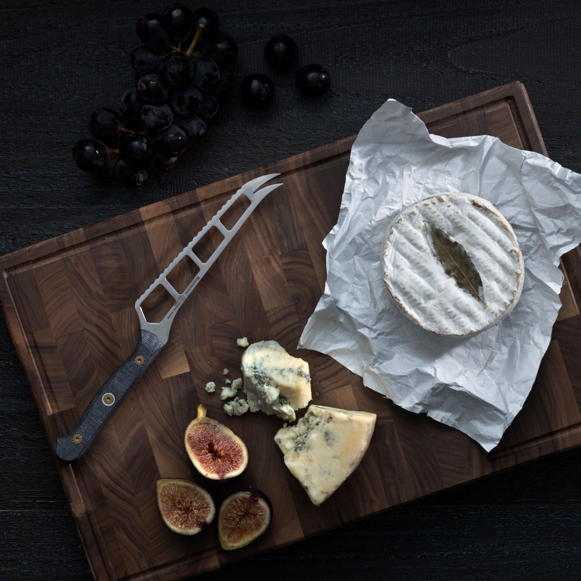 Messermeister Custom Cheese & Tomato Knife on a cutting board with cheese and figs that lay on a dark wood surface with grapes on it. 