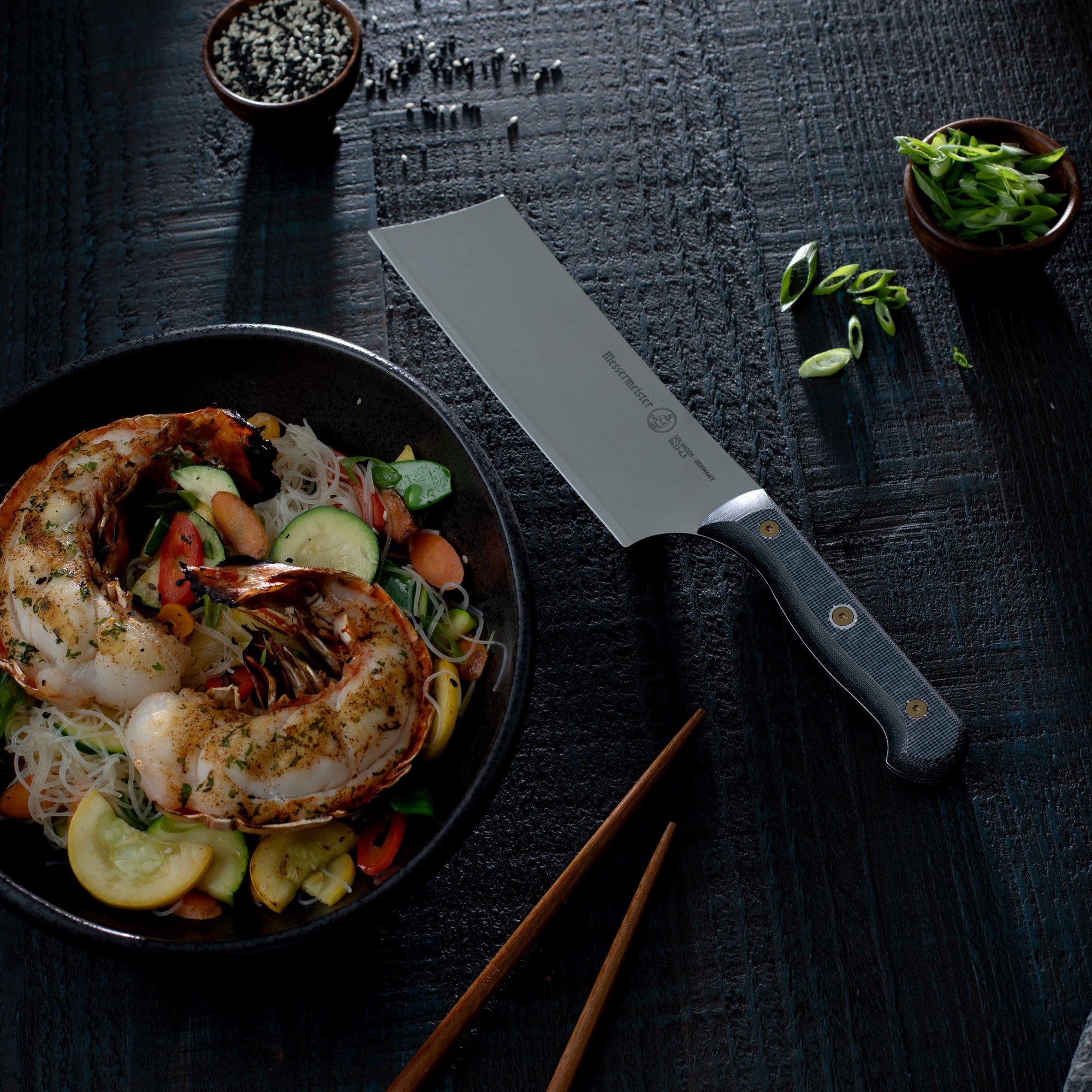 Plated dish with seafood and vegetables, Messermeister Custom Nakiri knife, and chopsticks on a dark surface