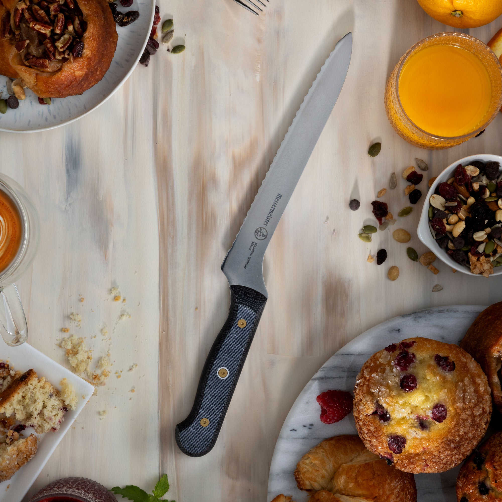 Messermeister Custom Offset Scalloped Bread Knife on a wooden surface with pastries, fruits, and a glass of orange juice.