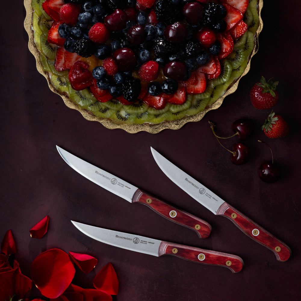 Messermeister Custom Red Steak Knives with fruit tart, cherries, strawberries and rose petals on a dark red surface