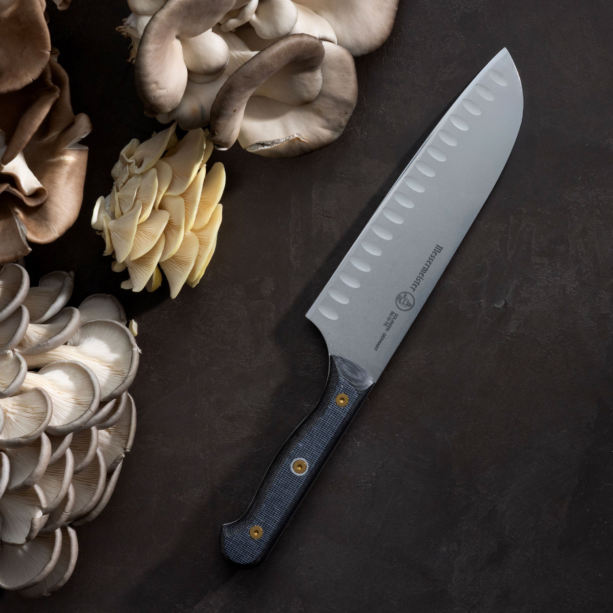 Messermeister Custom Santoku with a black micarta handle with gourmet mushrooms on a dark brown surface