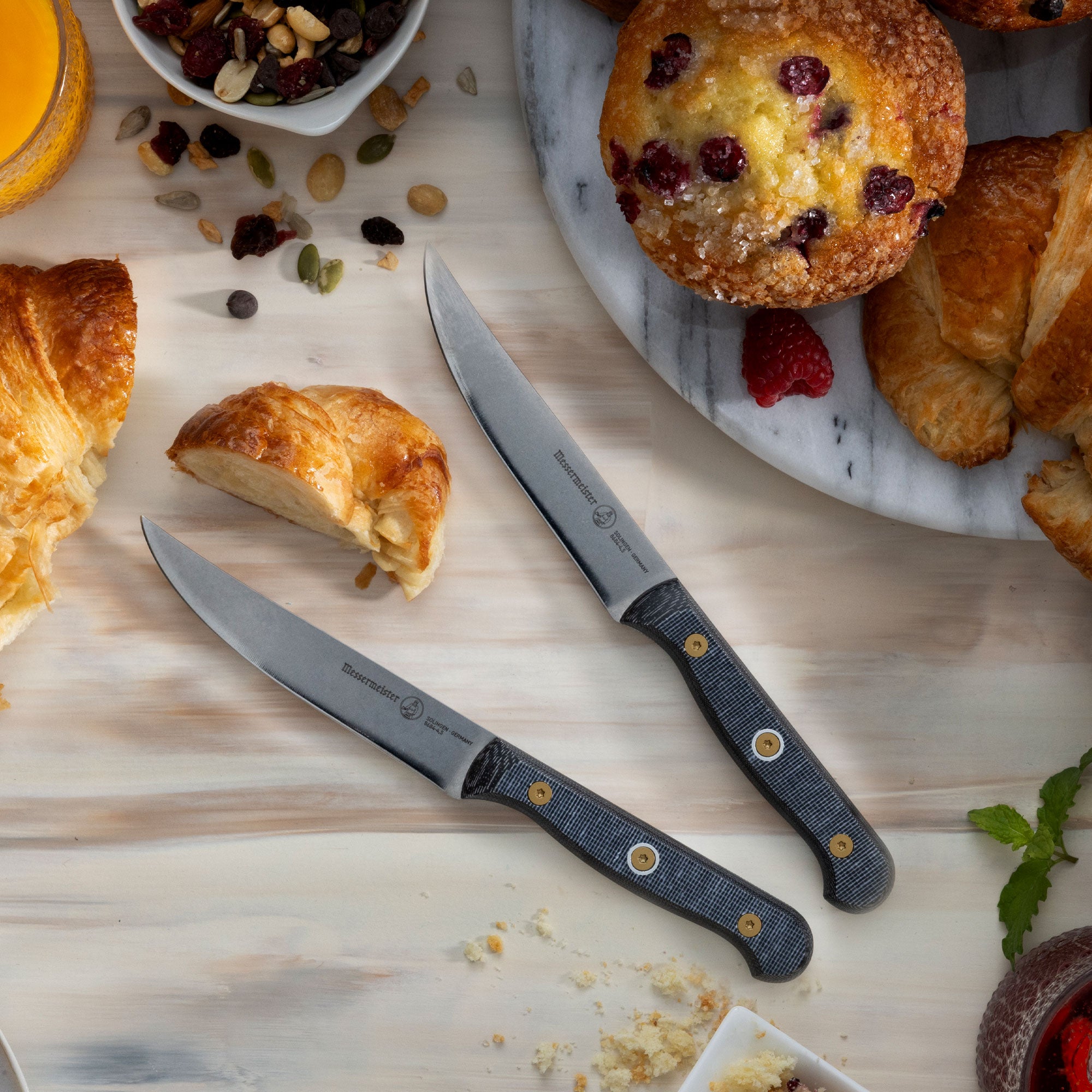 Messermeister Custom Steak Knives with black micarta handles in cafe setting with pastries on a light wood surface