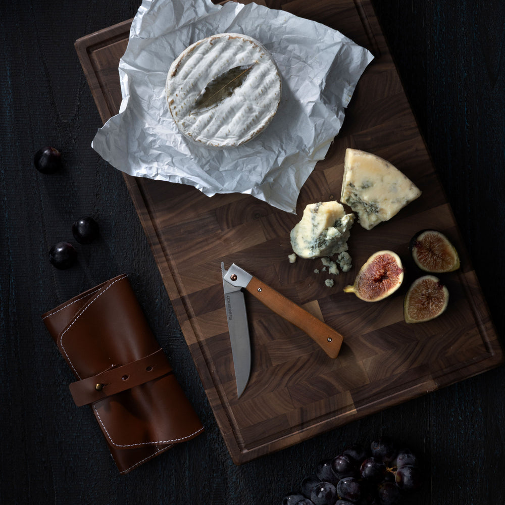 Messermeister Folding Steak Knife on a cutting board with cheese and figs that lay on a dark wood surface with a leather roll and grapes on it. 