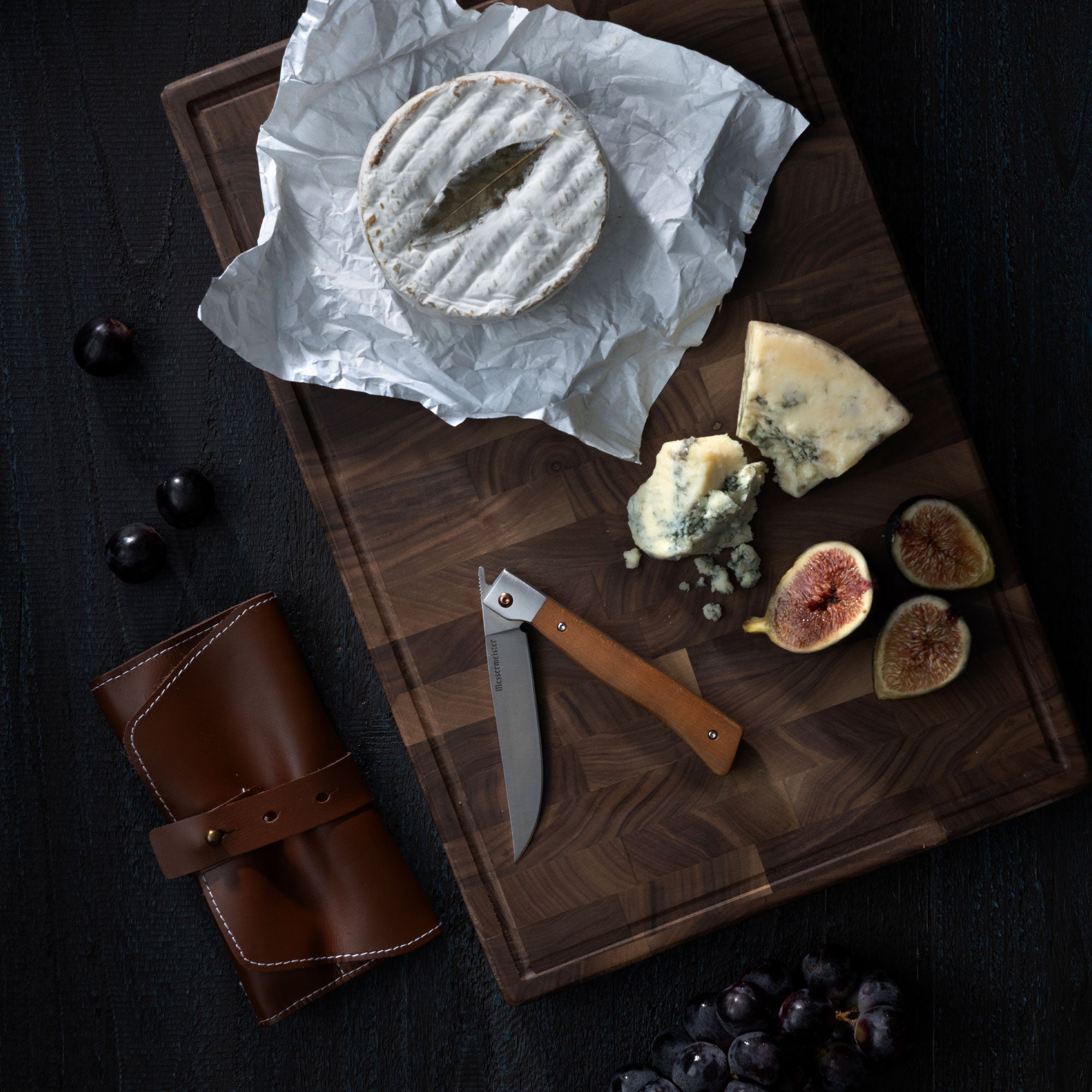 Messermeister Folding Steak Knife on a cutting board with cheese and figs that lay on a dark wood surface with a leather roll and grapes on it. 