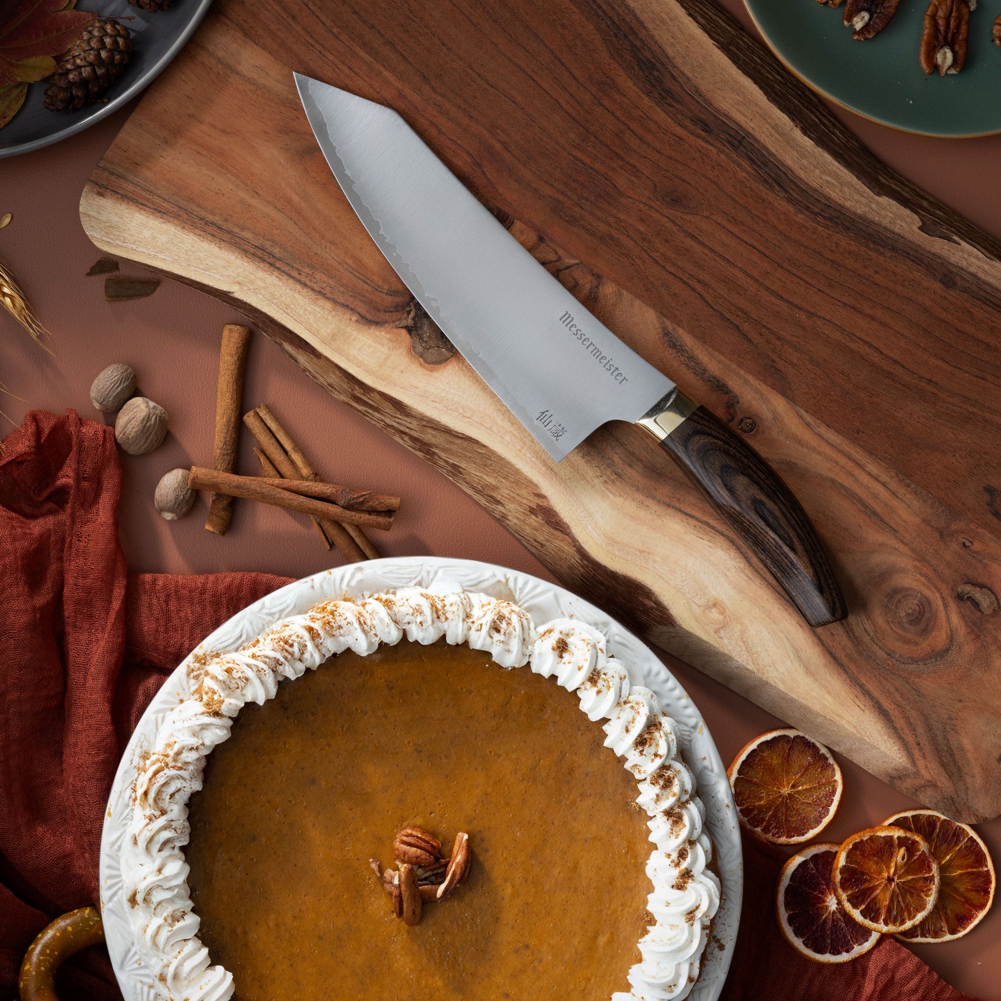 Messermeister Kawashima on Wood Cutting Board with pumpkin pie, cinnamon sticks, dried oranges, and a dark orange linen on a terracotta color surface.
