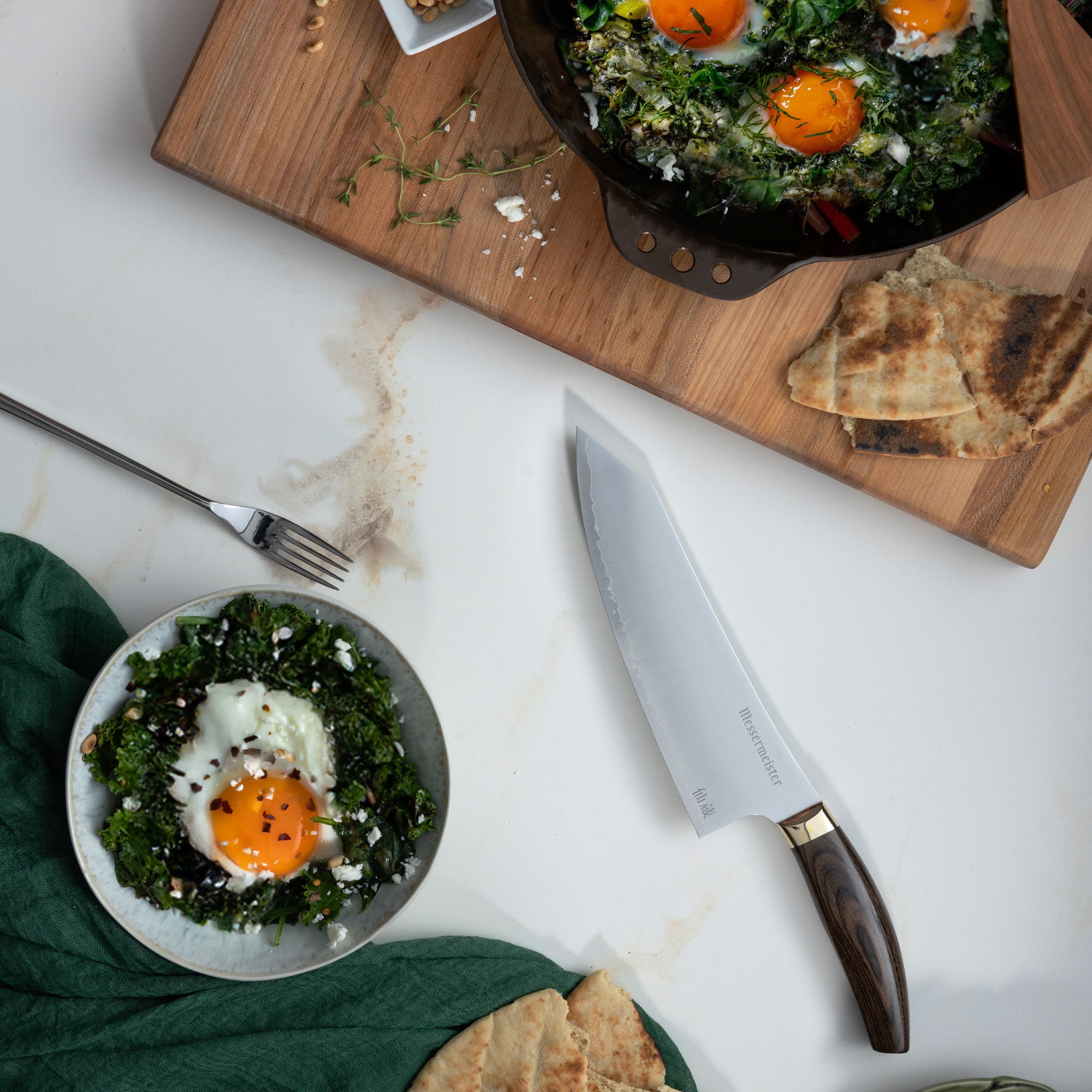 Messermeister Kawashima Chef's Knife with Mother's Day egg green shakshuka in a skillet and a bowl that is laying on a white marble countertop with a green linen and fork