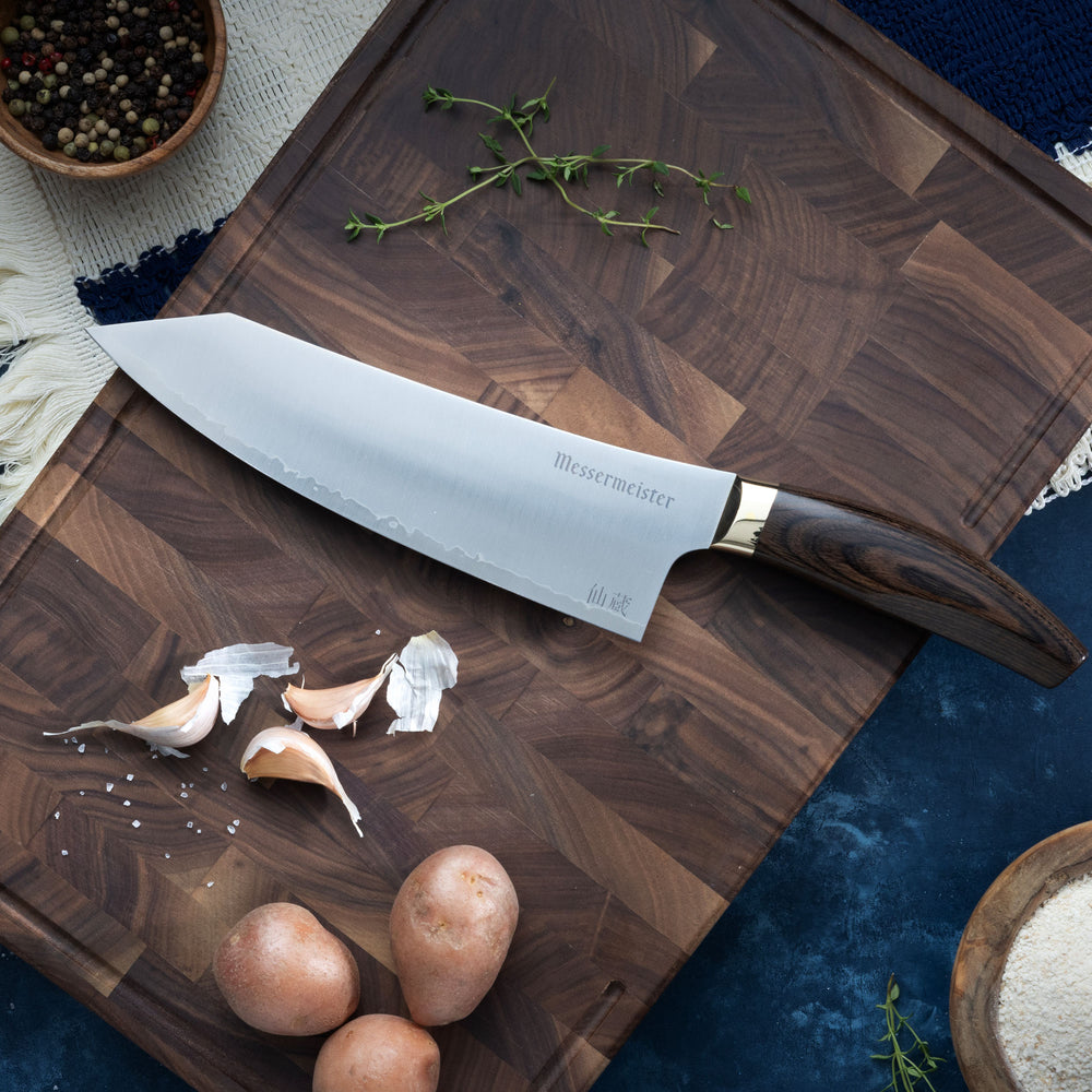 Messermeister Kawashima Chef Knife with potatoes and garlic on a cutting board that is laying on a blue surface