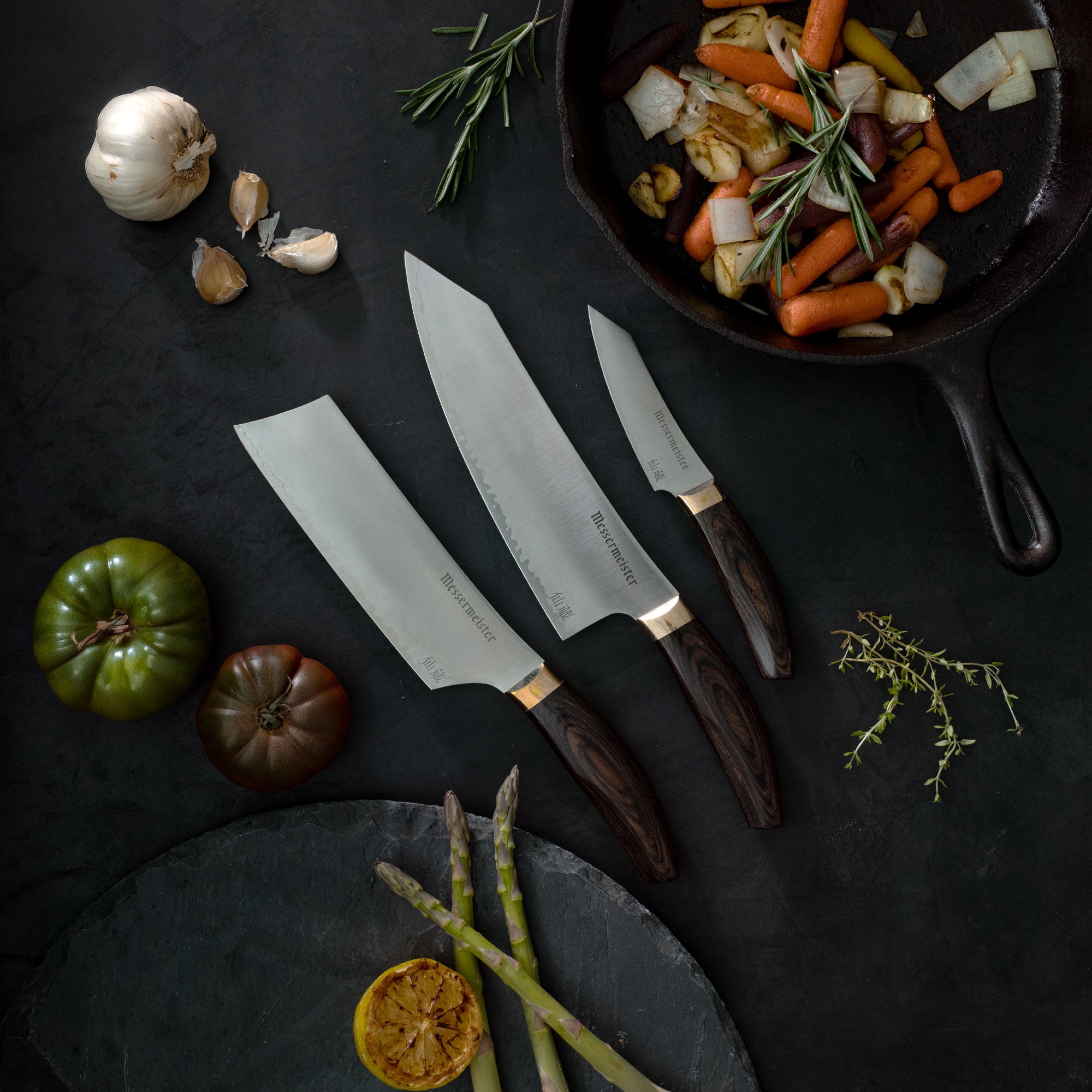 Messermeister Kawashima Nakiri, Chef's and Paring knives on a dark surface with vegetables and a pan of food.