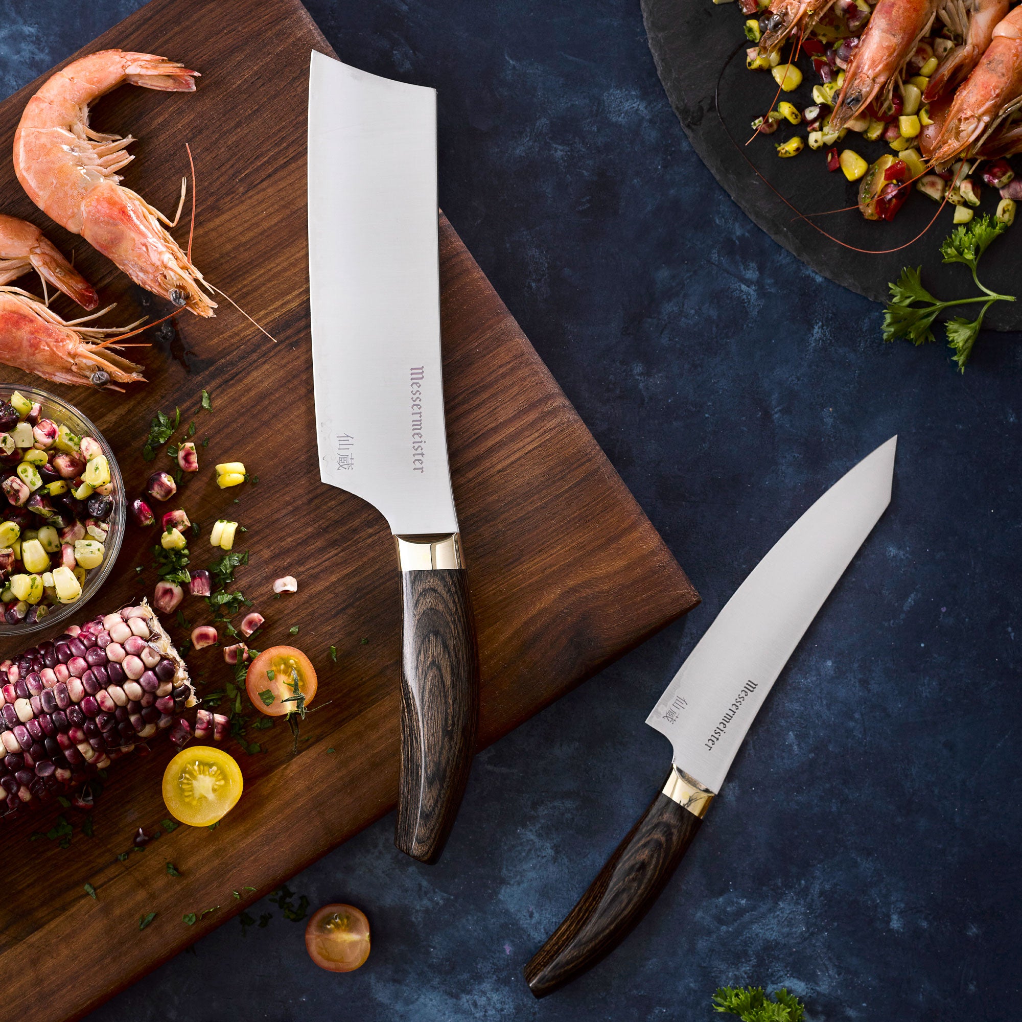 Messermeister Kawashima Nakiri and Utility Knife on cutting board with corn and prawns on a dark blue surface