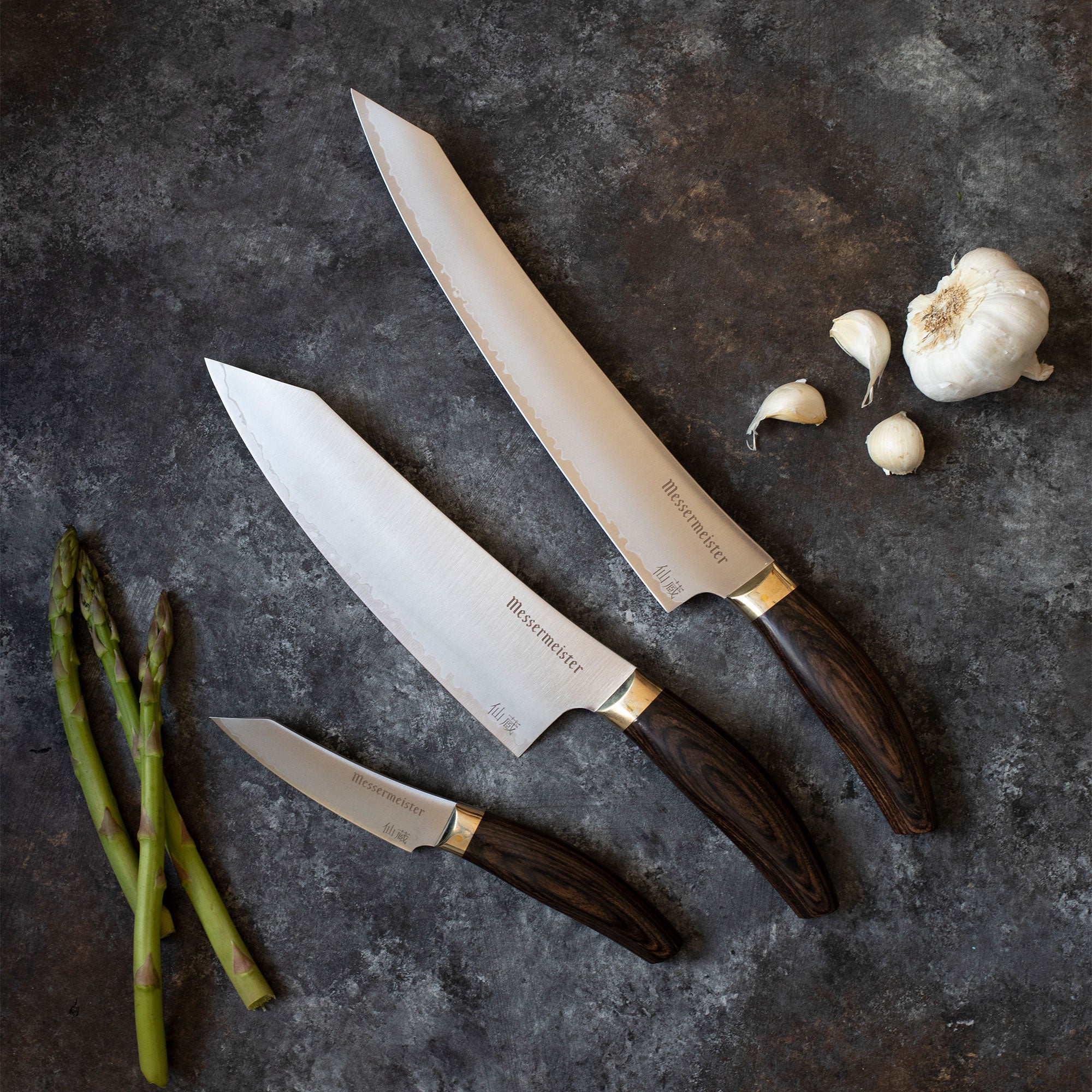 Messermeister Kawashima Slicer, Chef's Knife, Paring Knife with asparagus and garlic on a dark gray and white surface.