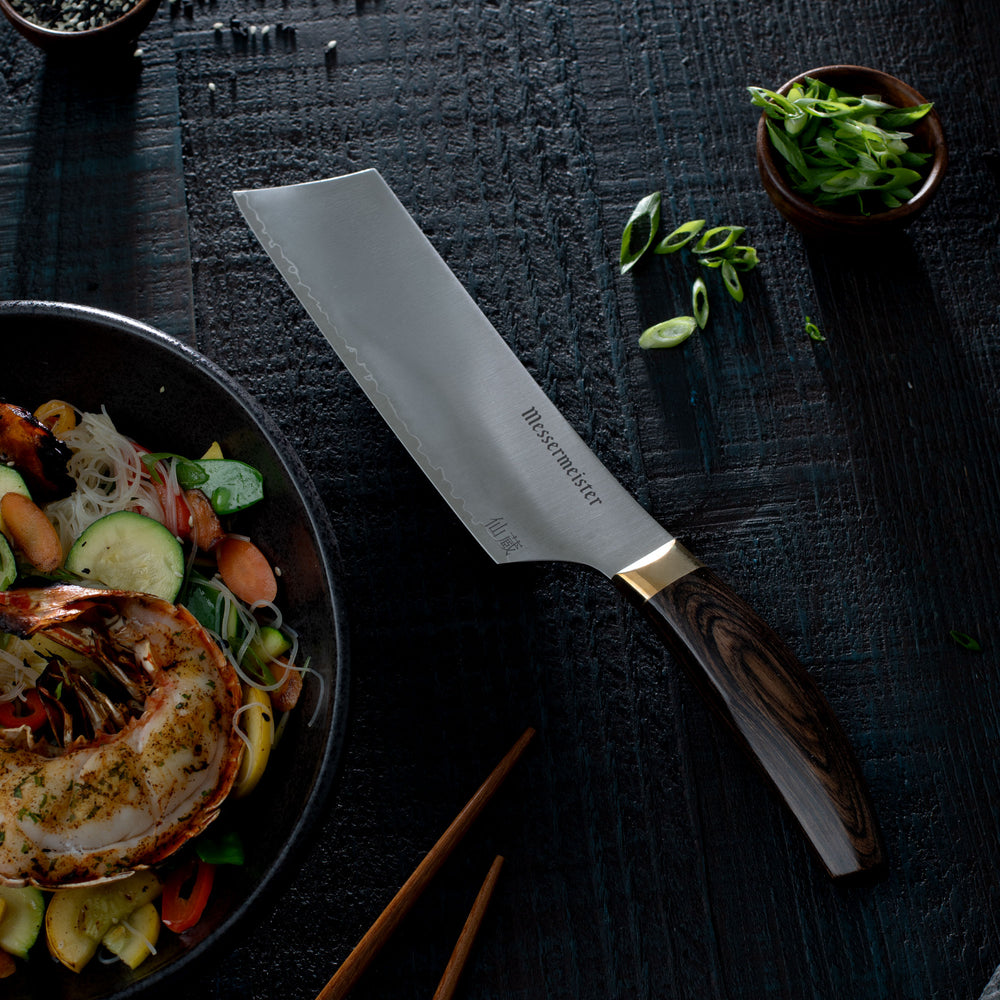 Messermeister Kawashima Nakiri Knife on a dark surface next to a bowl of stiry fry noodles and lobster tail and chopsticks.