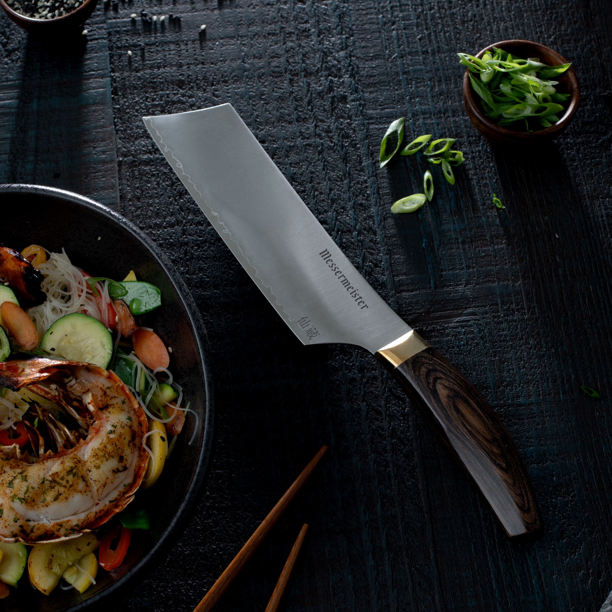 Messermeister Kawashima Nakiri Knife on a dark surface next to a bowl of stiry fry noodles and lobster tail and chopsticks.