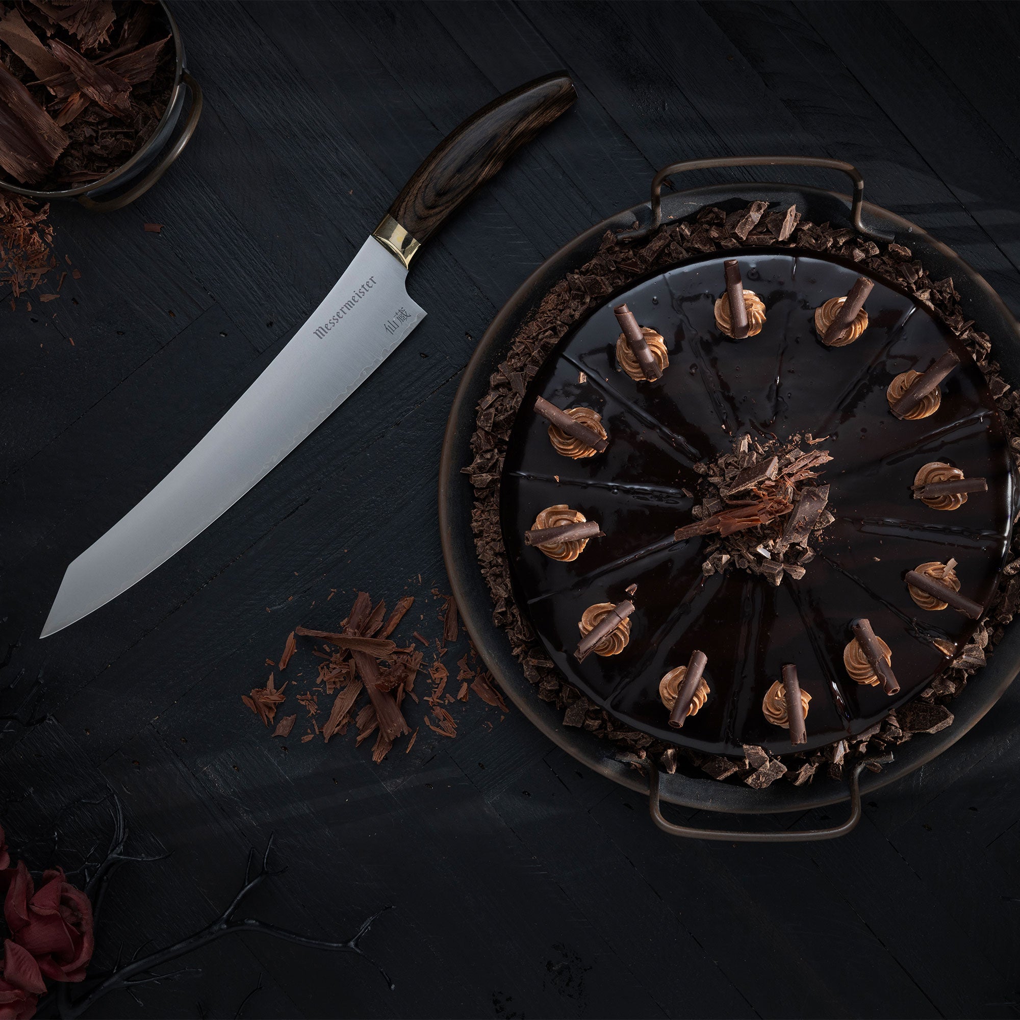 Messermeister Kawashima Slicing Knife laying besides a dark chocolate cake on a black wood surface.