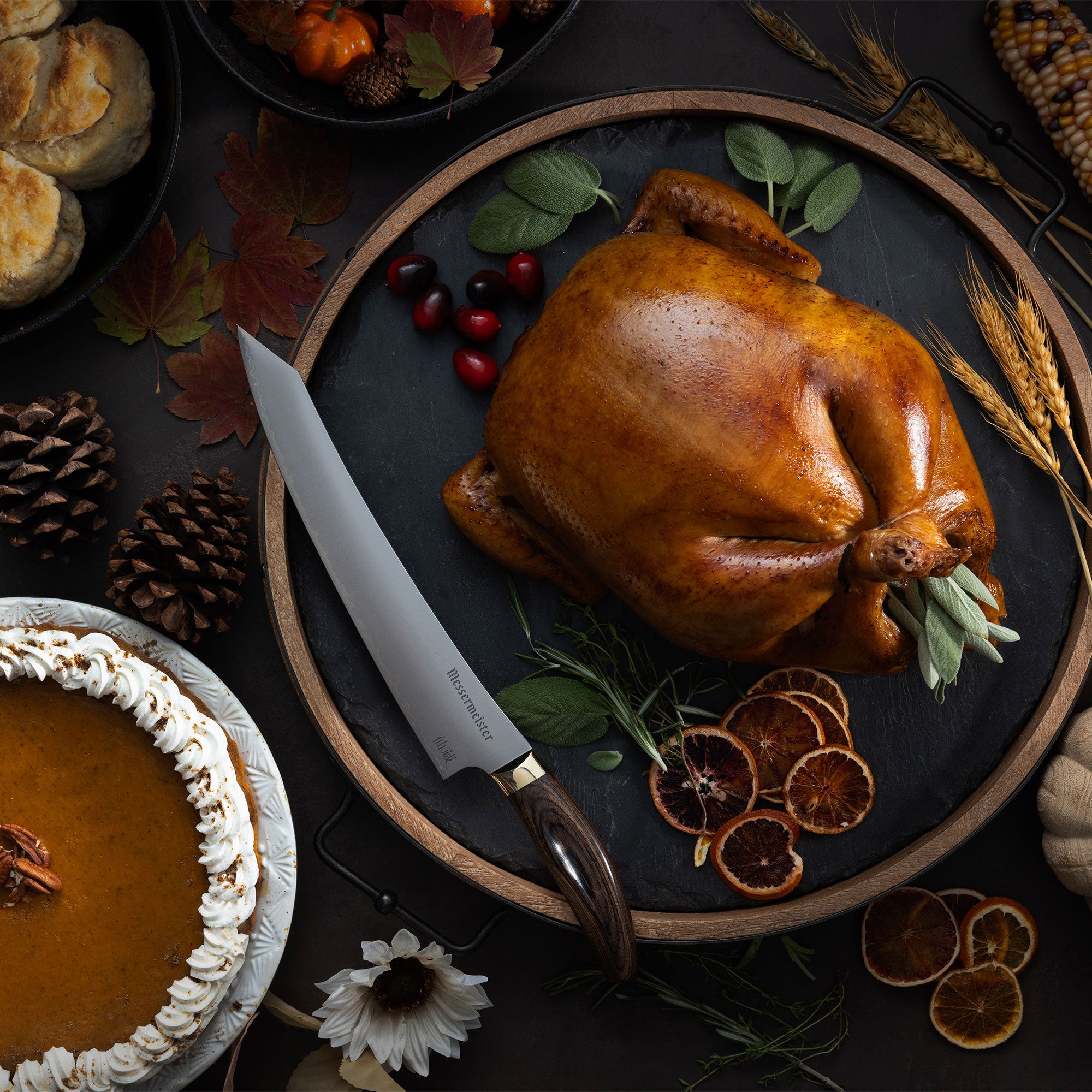 Messermeister Kawashima Slicing Knife on a plate with a Thanksgiving turkey, slices of orange, and cranberries that is surrounded by fall decor on a dark surface